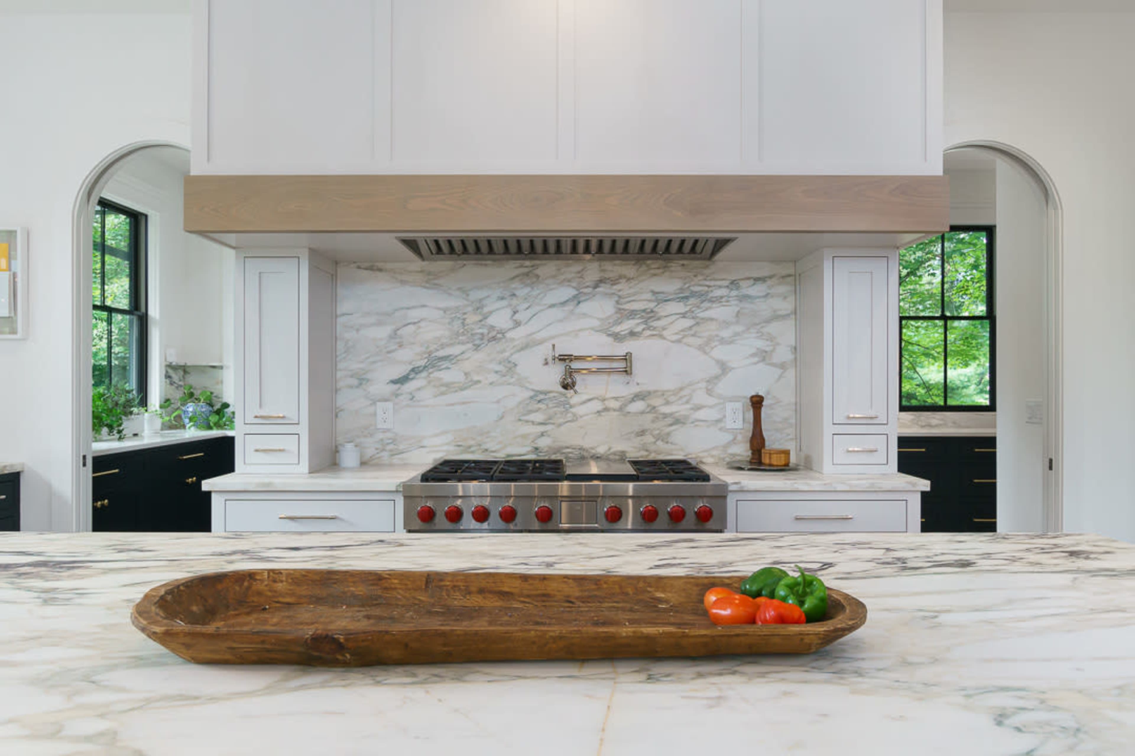 A modern kitchen featuring a marble backsplash, a professional-grade stovetop with red knobs, and a wooden tray with tomatoes and bell peppers on a marble countertop.