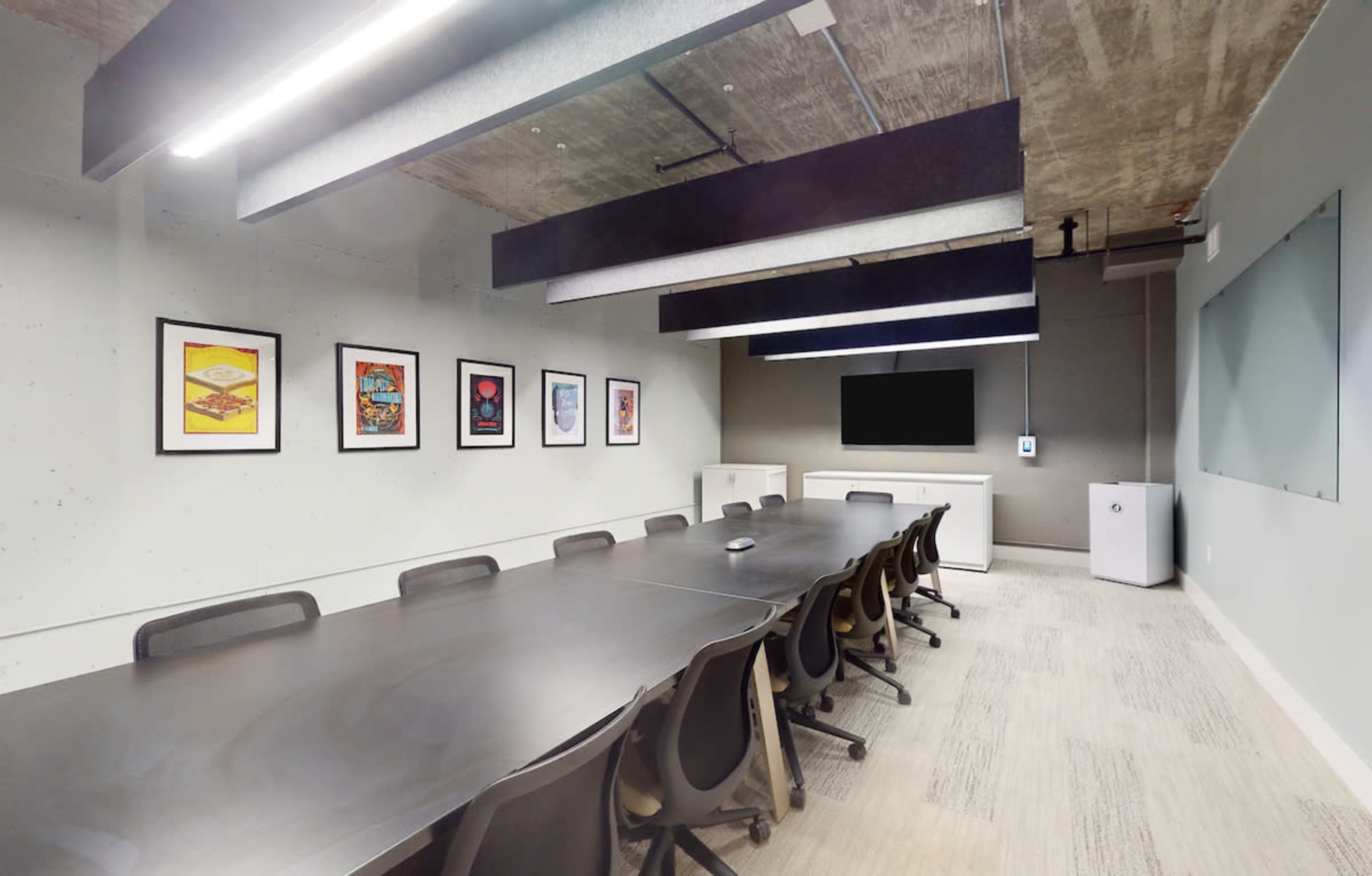 A modern conference room features a long table surrounded by chairs, with framed artwork on the walls and a large screen for presentations.