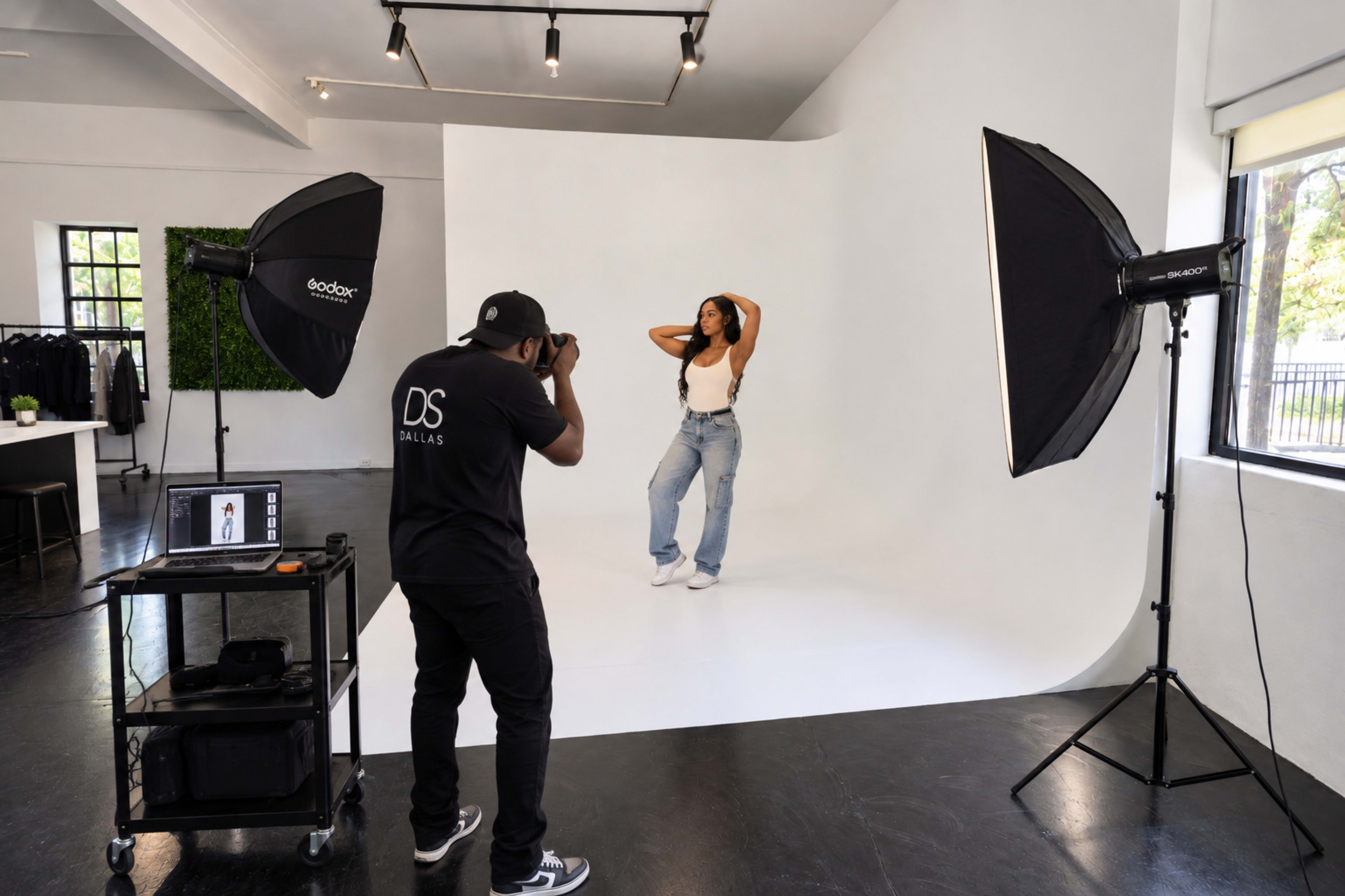 A photographer captures an image of a model posing against a white backdrop in a studio setting equipped with lighting equipment.