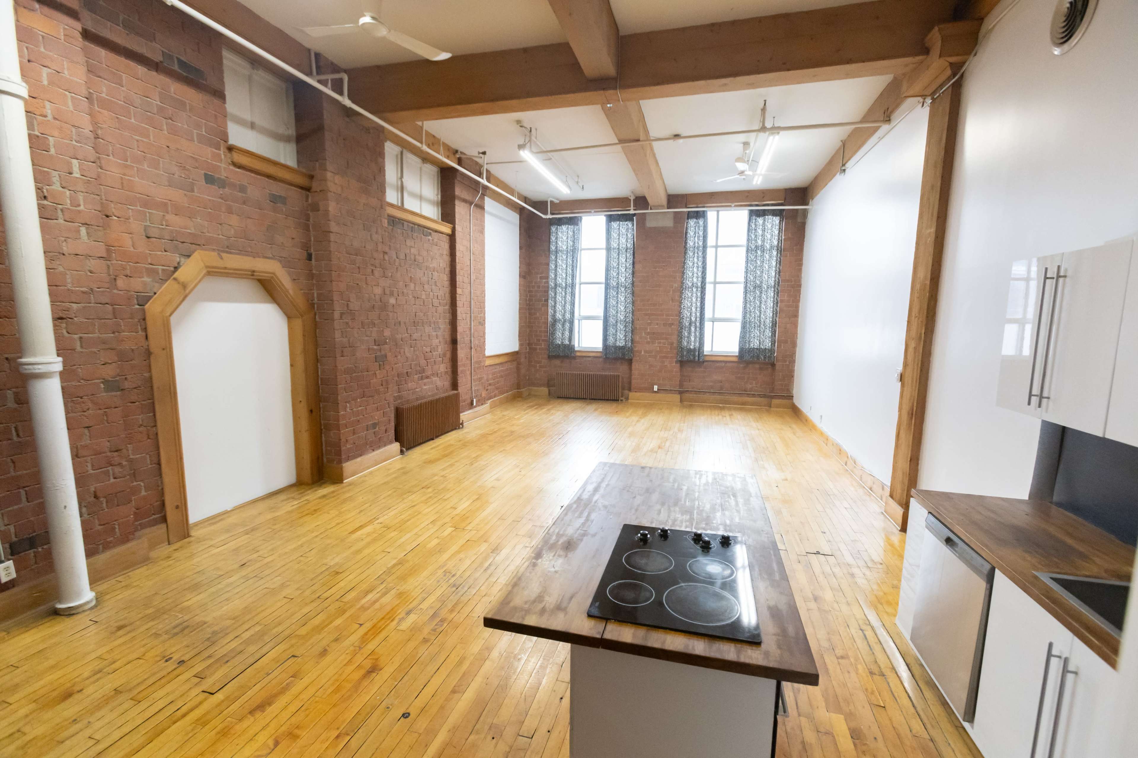 The image shows an empty loft-style room with brick walls, large windows, and an island with a cooktop in the center.