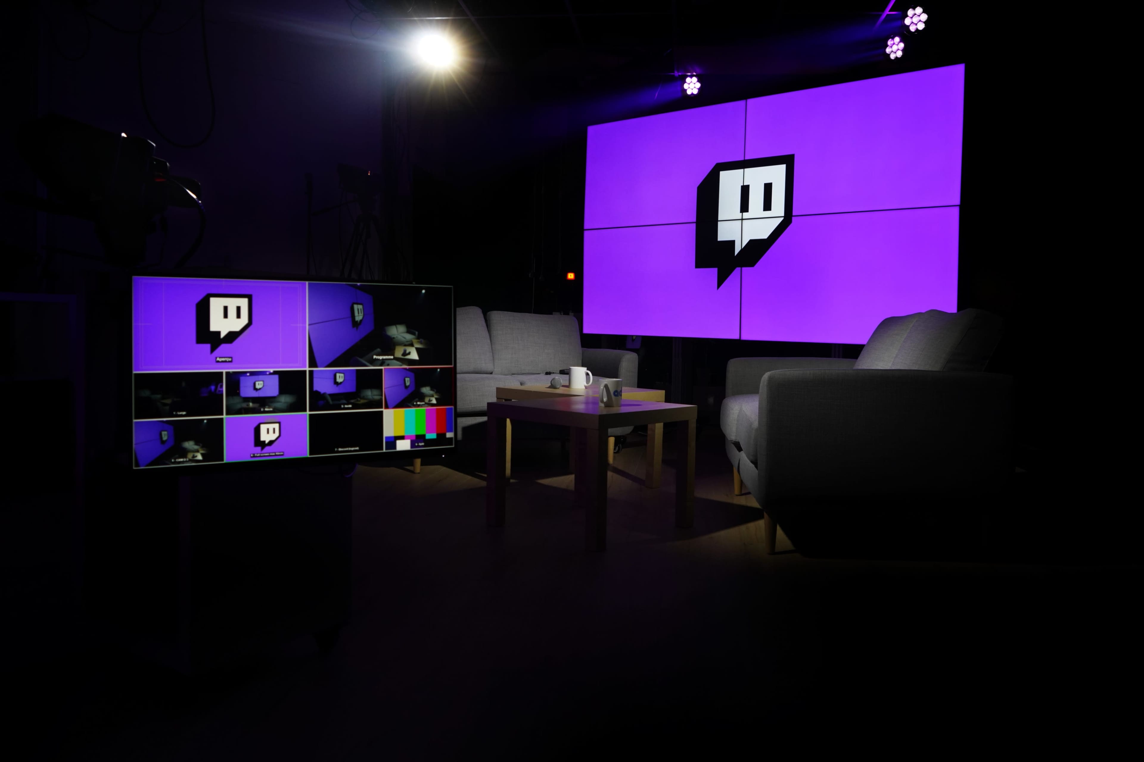 A modern streaming setup with a large purple Twitch logo displayed on a screen, alongside a smaller monitor showing various streaming feeds, two armchairs, and a coffee table.
