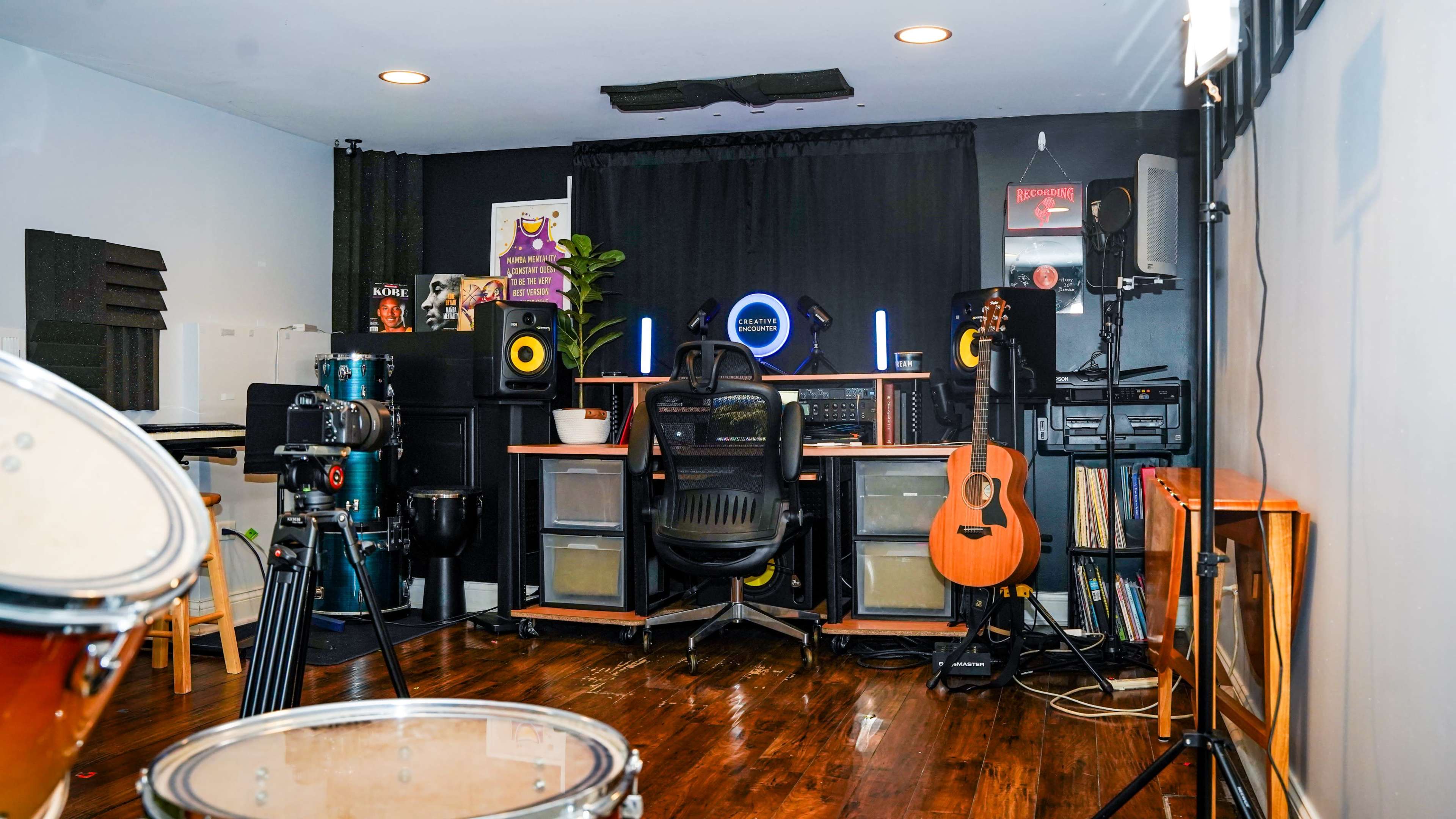 A music studio setup with a desk, computer equipment, a guitar, and a drum kit visible in the foreground.