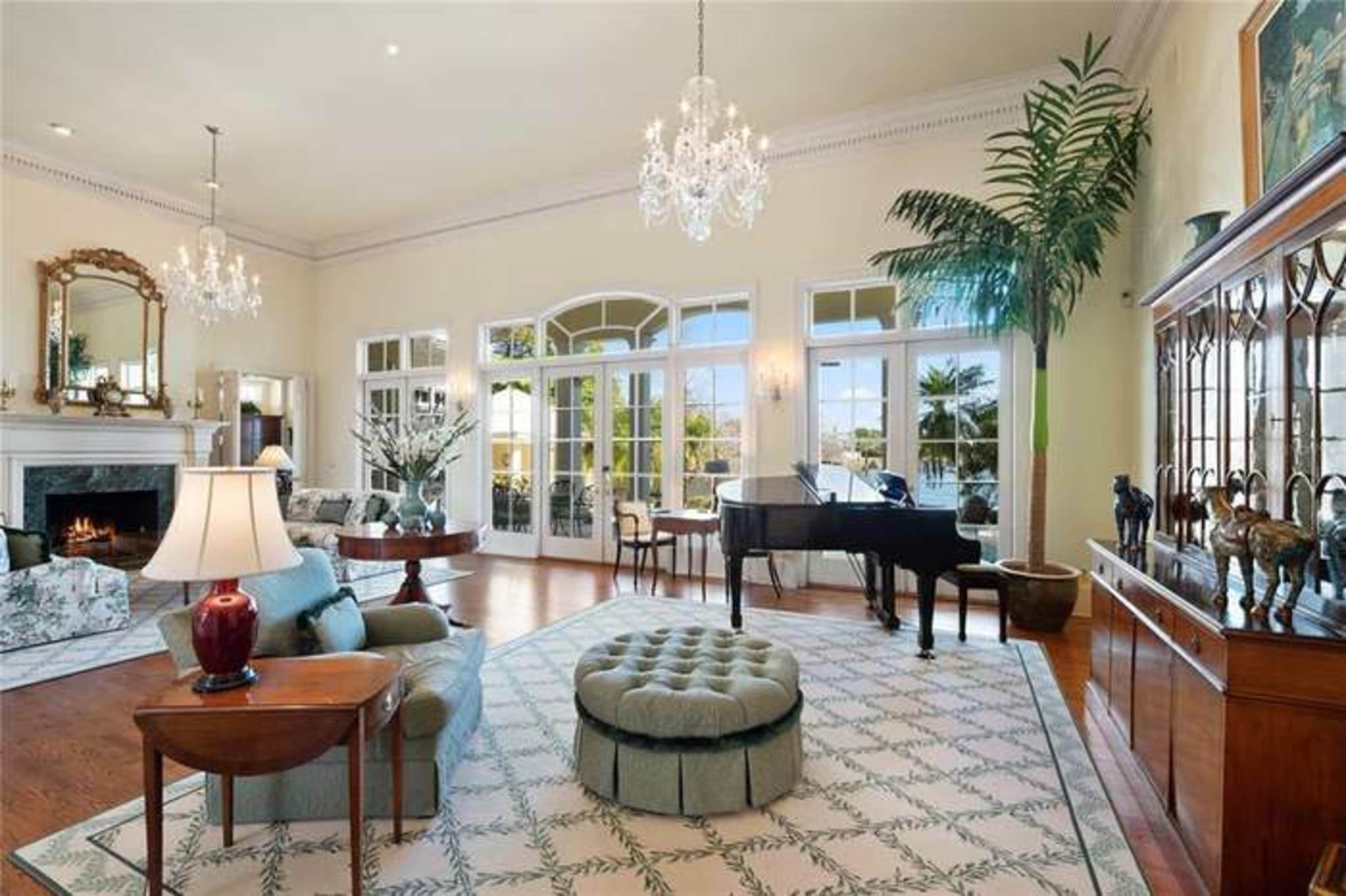 The spacious living room features large windows, a grand piano, and elegant furnishings arranged around a central rug.