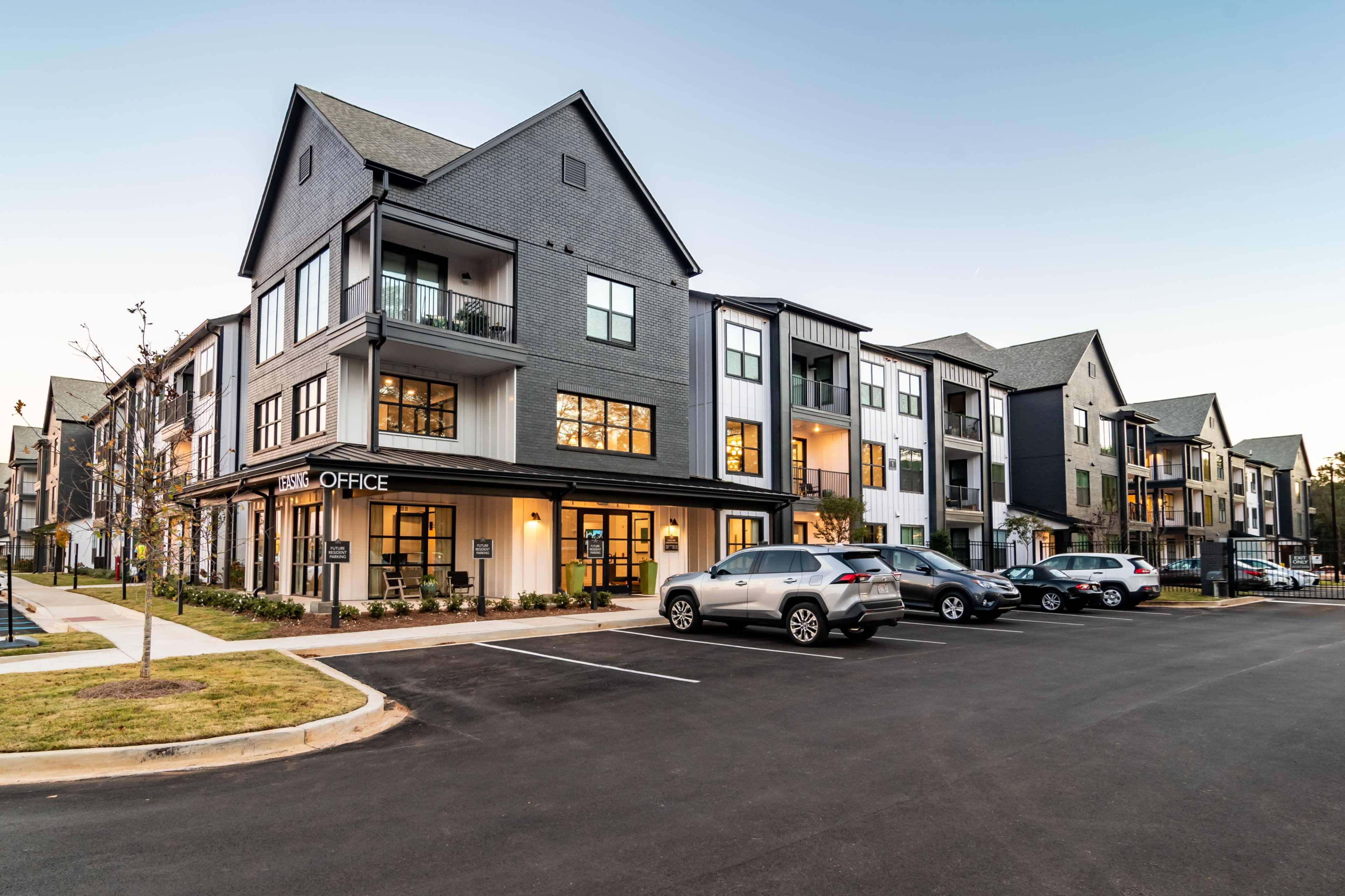 A modern multi-unit apartment building features a leasing office on the ground floor and several vehicles parked in front.
