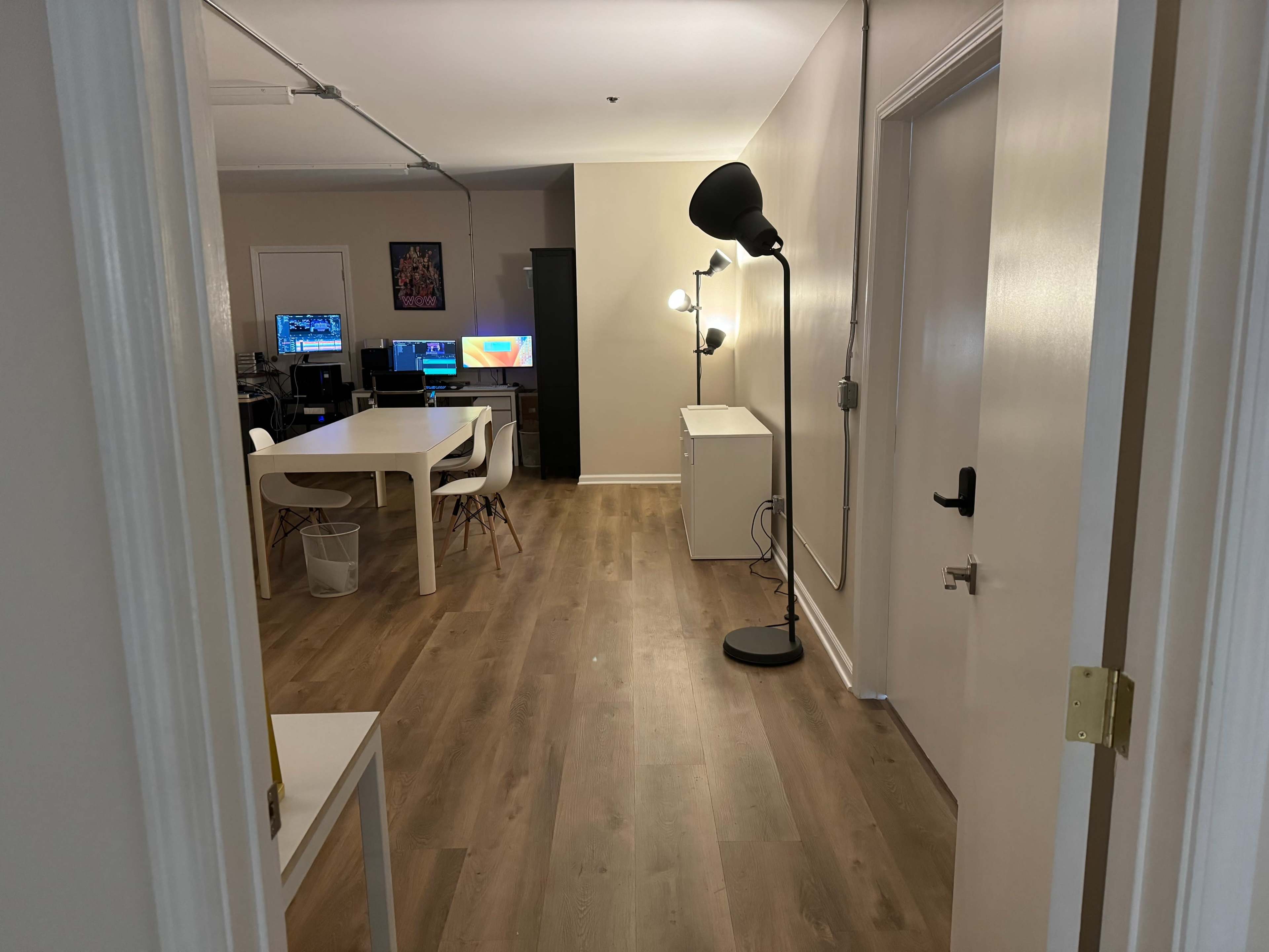 The image shows a hallway leading to a room with a white table and chairs, a black lamp, and two computers in the background.