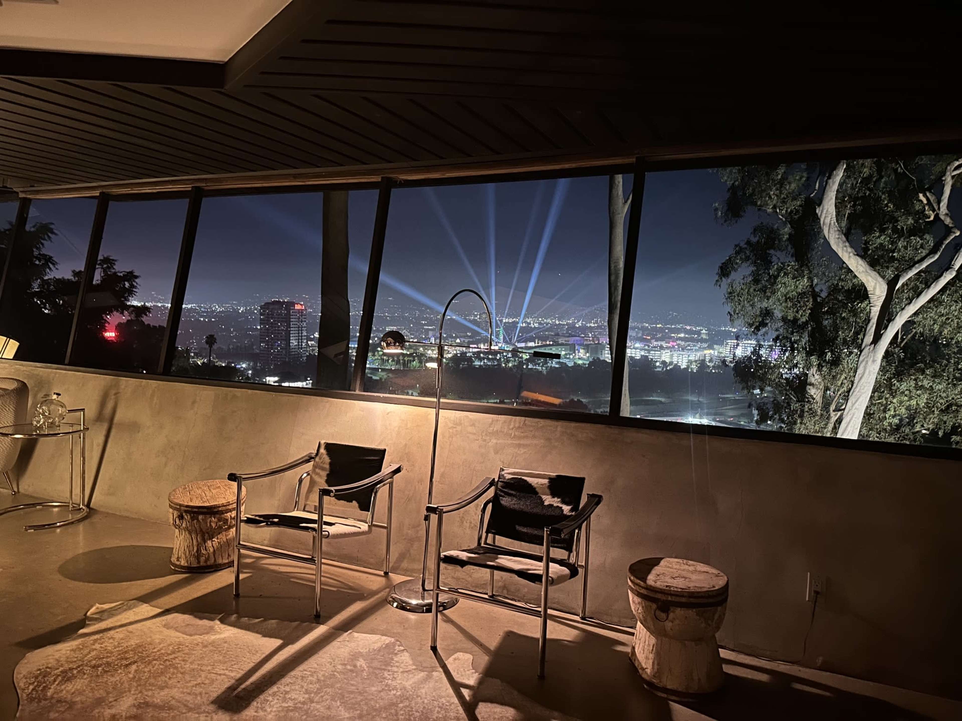 The image shows a modern interior with two chairs and a lamp, overlooking a city skyline illuminated by beams of light in the night sky.