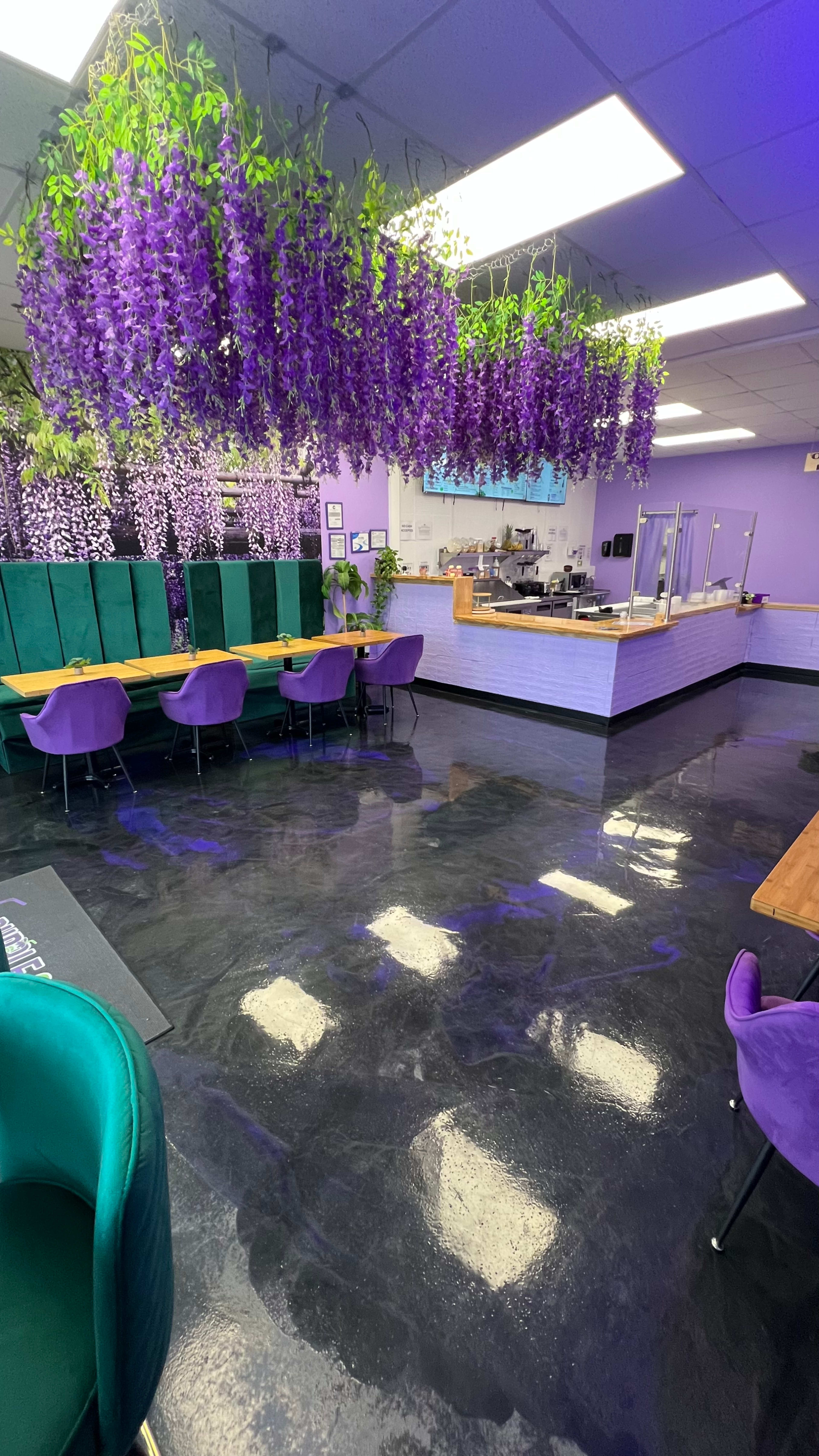 The image shows a café interior featuring purple flowers hanging from the ceiling, purple seating, and a counter area at the back.