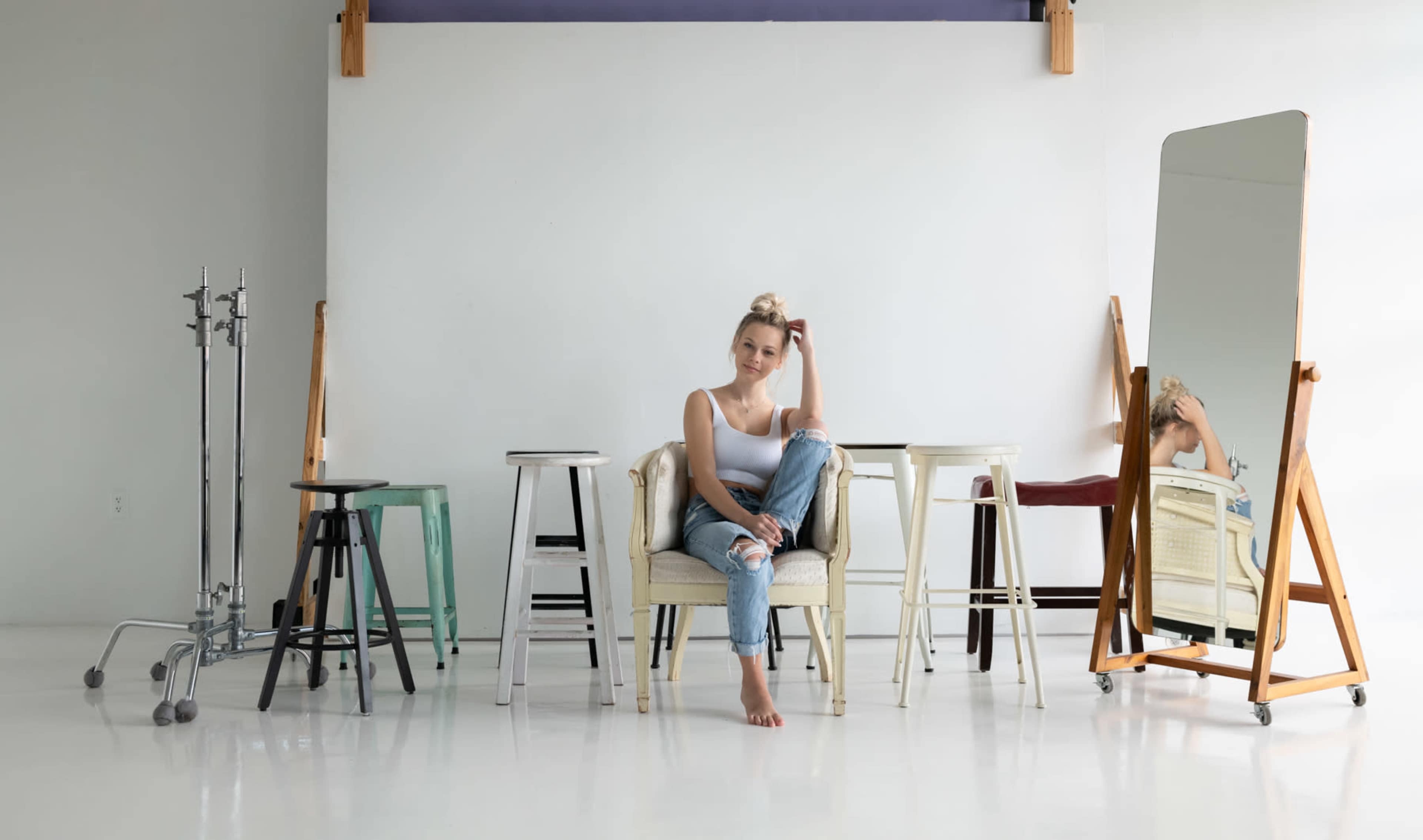 A young woman sits on a chair surrounded by various stools and a large mirror in a bright, minimalist studio space.