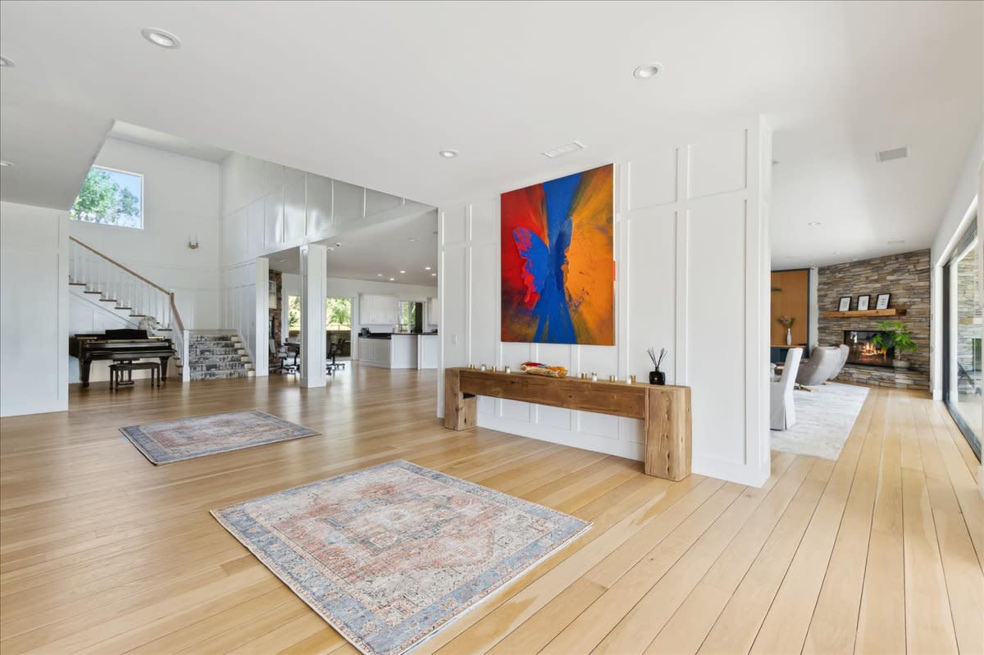 The scene shows a spacious, modern interior with wooden flooring, a large abstract painting on one wall, and a view of a living area with a fireplace and staircase visible in the background.