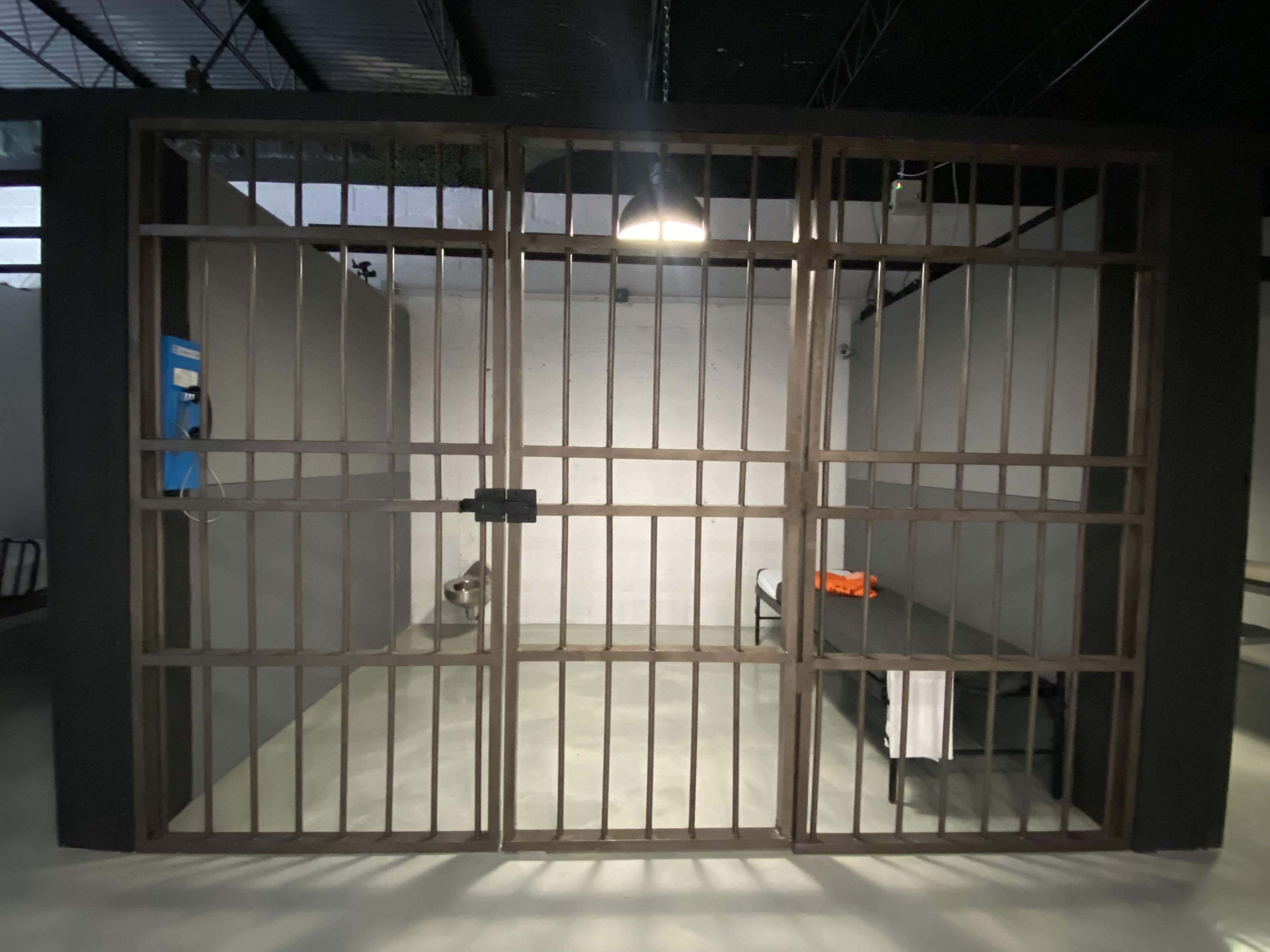 The image shows a locked jail cell with barred metal doors, a bench inside, and a wall-mounted payphone.