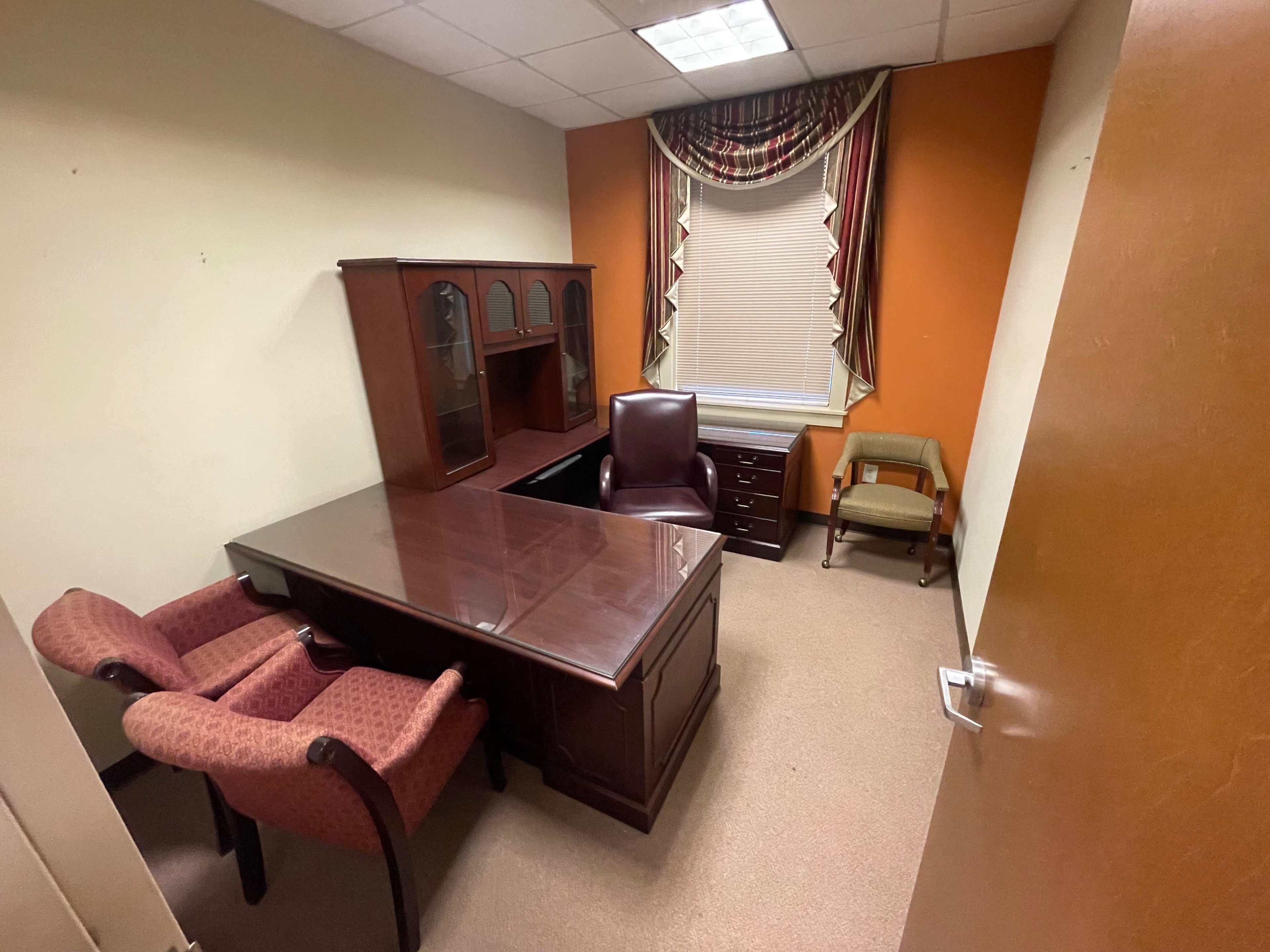 A small office with a wooden desk, two upholstered chairs, a filing cabinet, and a window covered by patterned drapes.