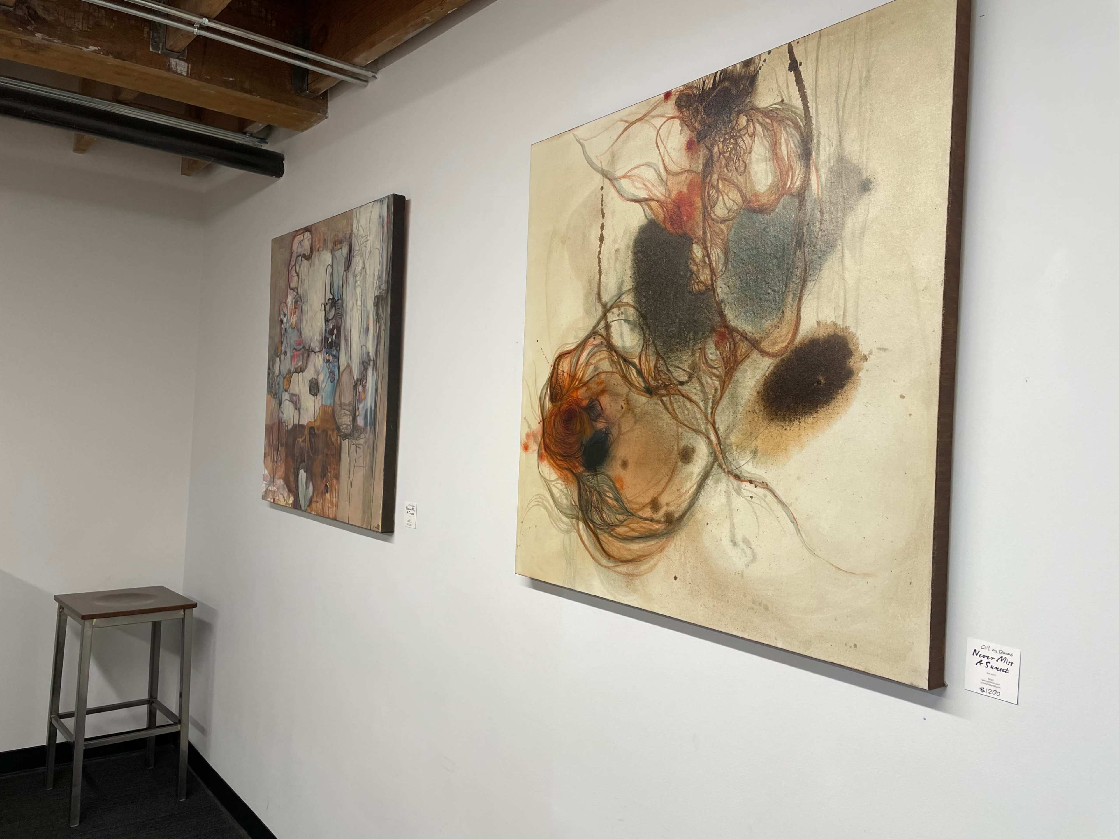 Two abstract paintings displayed on a white wall in an art gallery, with a metal stool positioned nearby.
