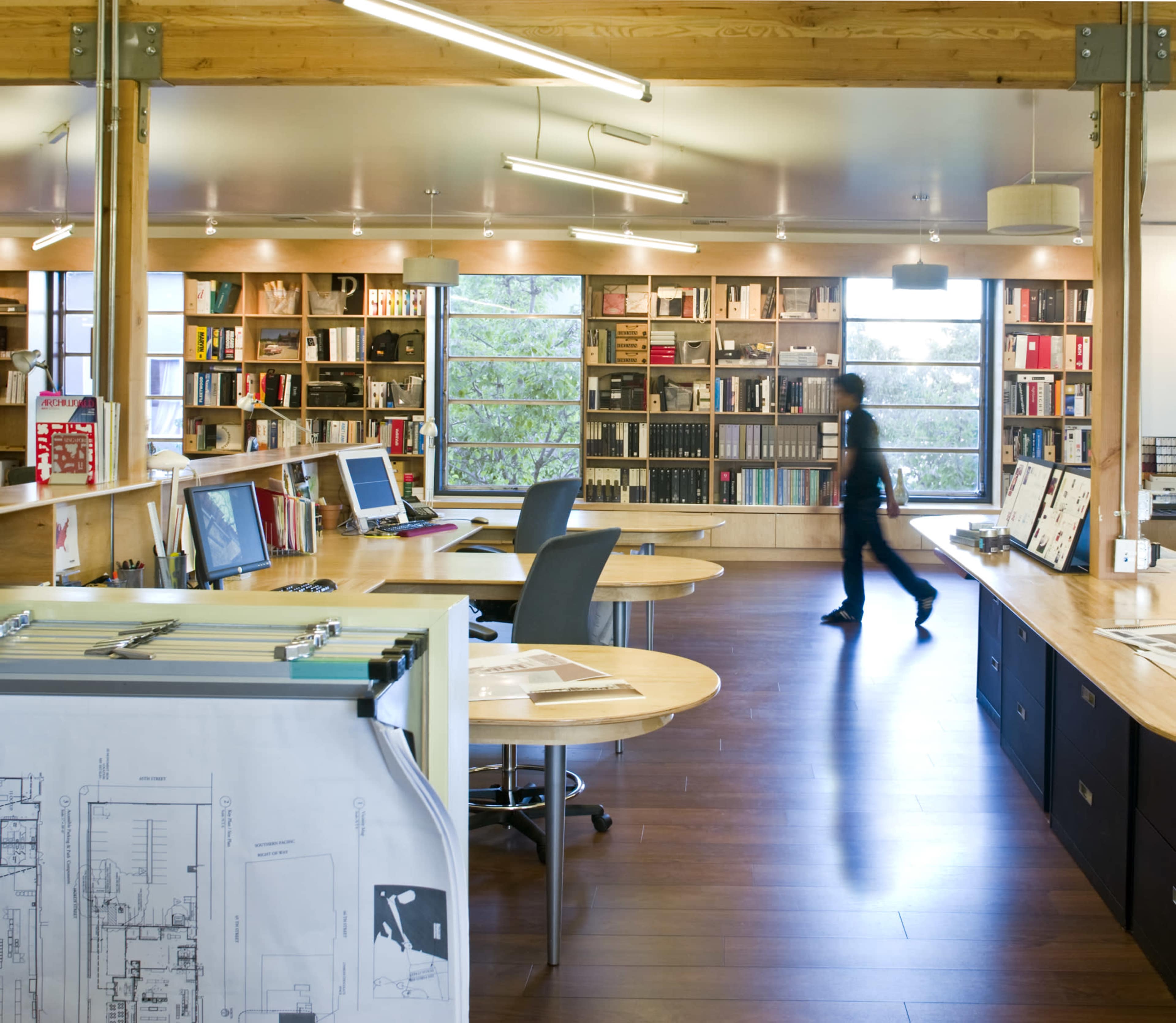 An office space features wooden desks with computers, shelves filled with books, and a person walking near a large window.