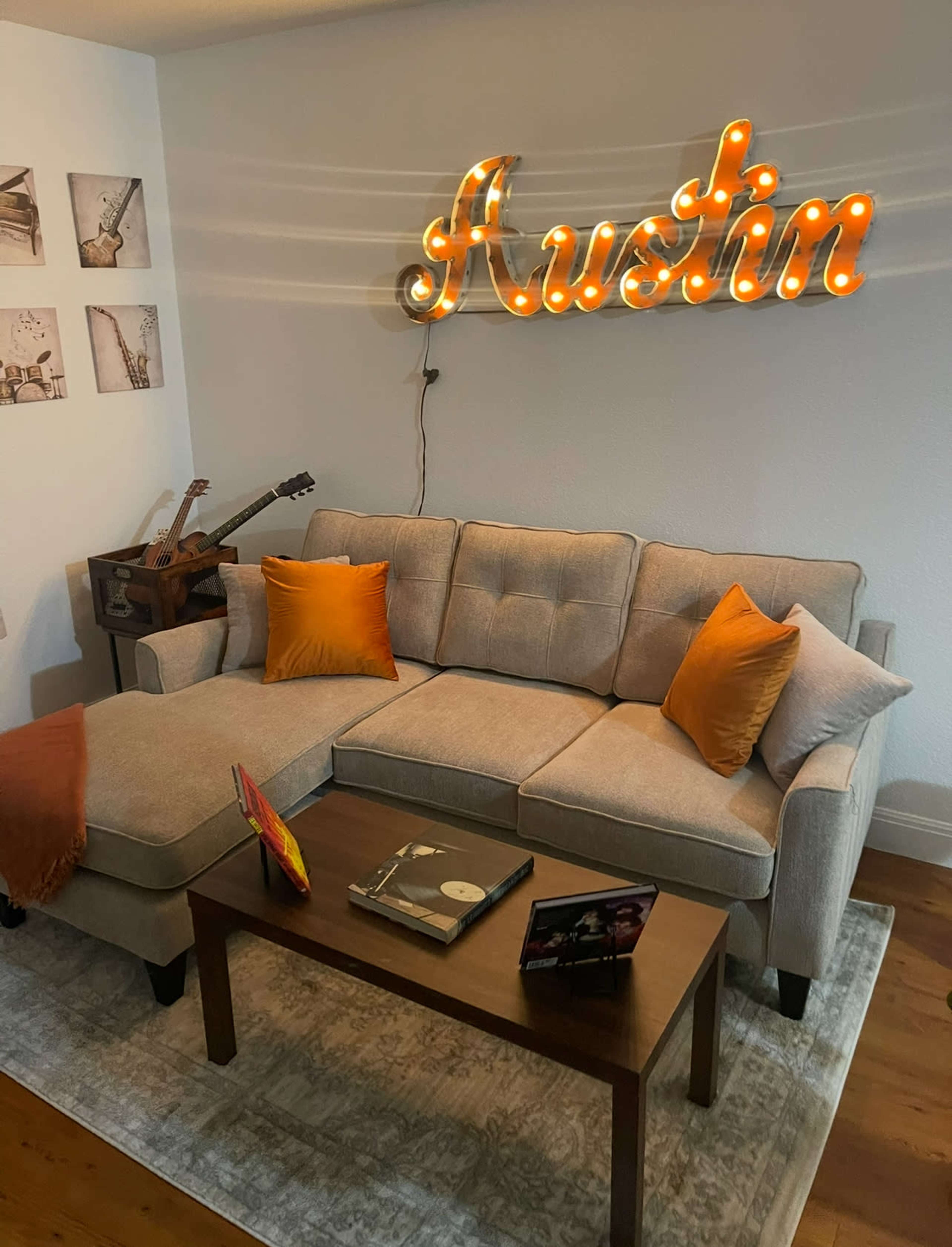 The image features a cozy living room with a gray sectional sofa adorned with orange pillows, a wooden coffee table, and a light-up sign that reads "Austin" on the wall.