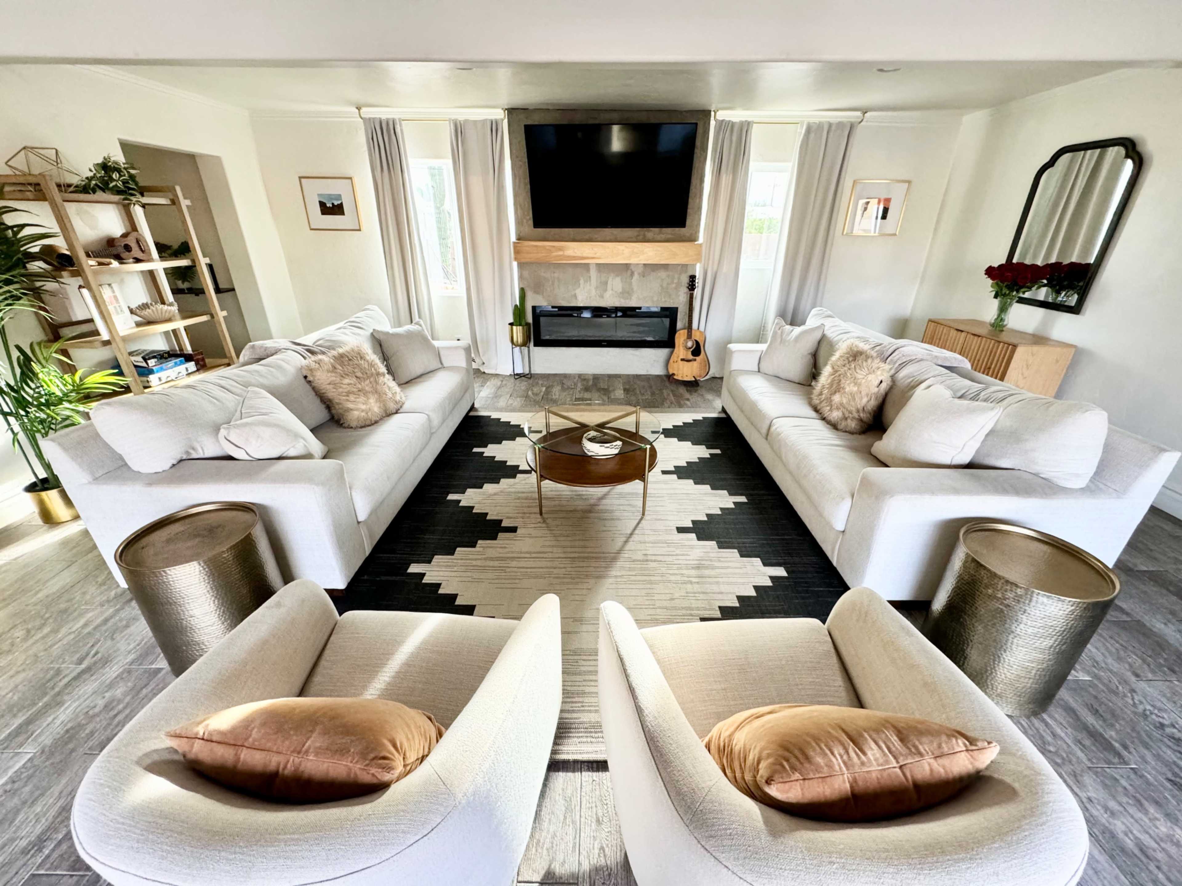 A modern living room features two large white sofas and two round side tables arranged around a central rug, with a fireplace and a television mounted on the wall.