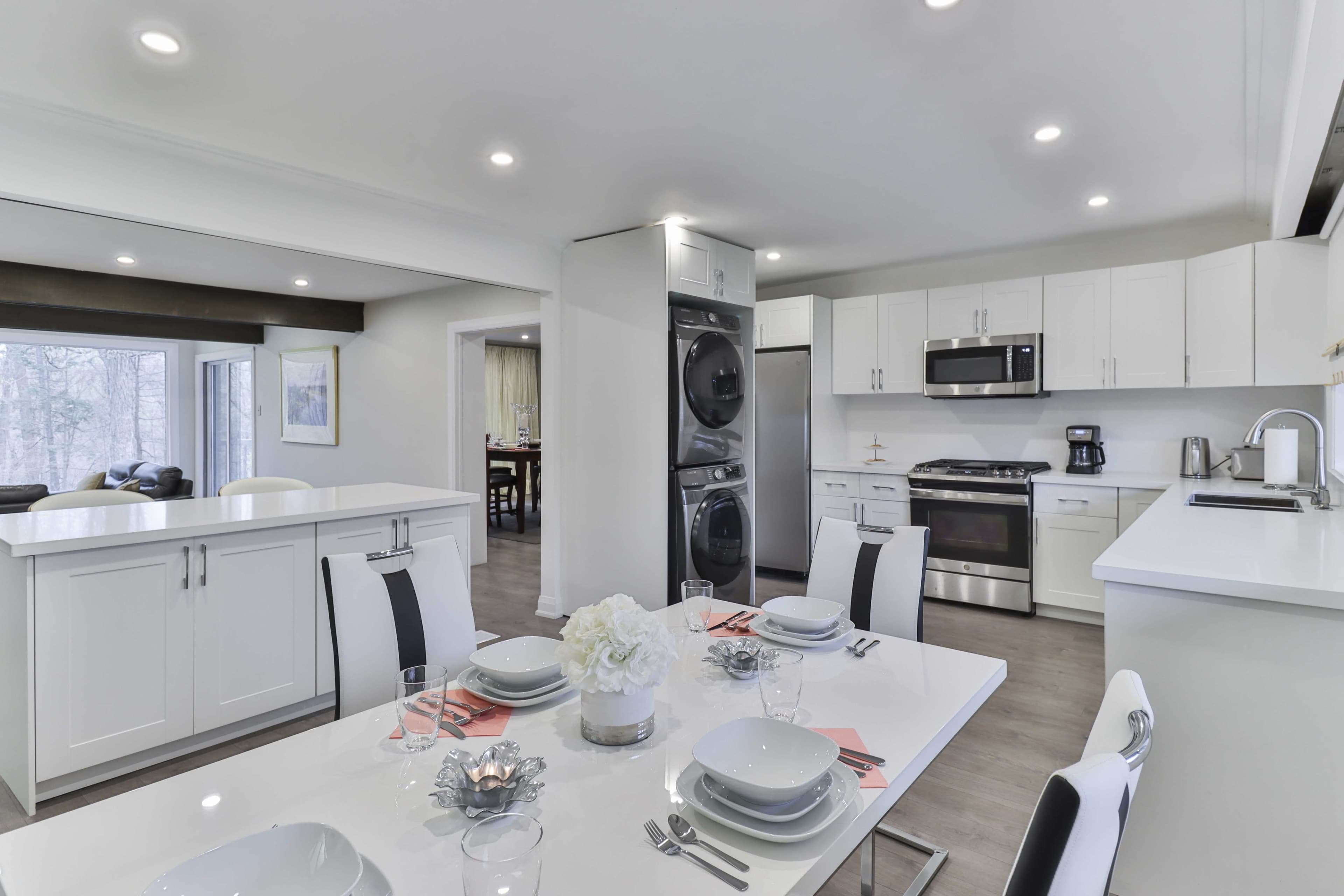 The image shows a modern kitchen and dining area featuring stainless steel appliances, a white table set for meals, and large windows allowing natural light.