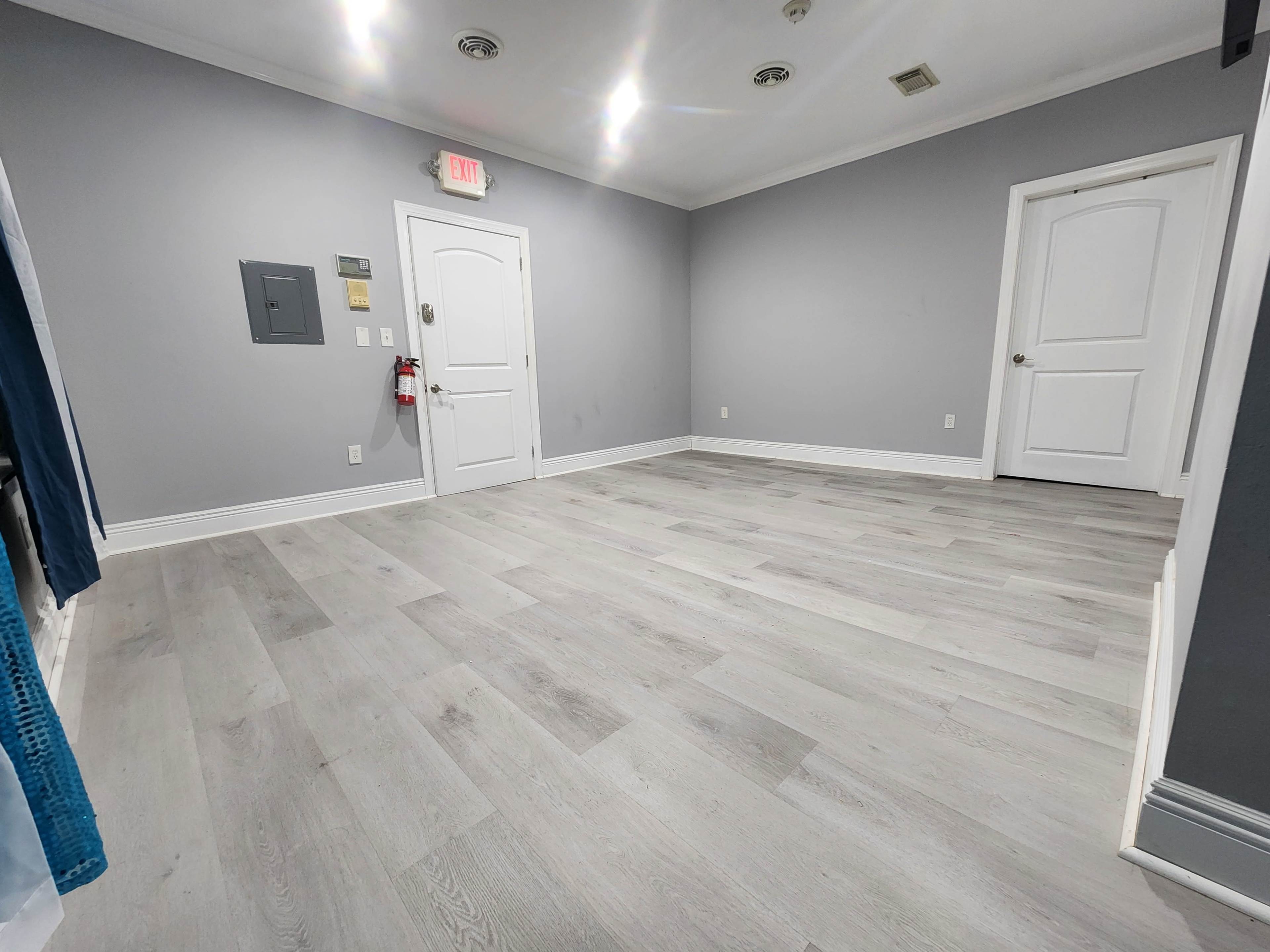 An empty room with light gray walls and a light wood floor, featuring a door and a small electrical panel on one wall.