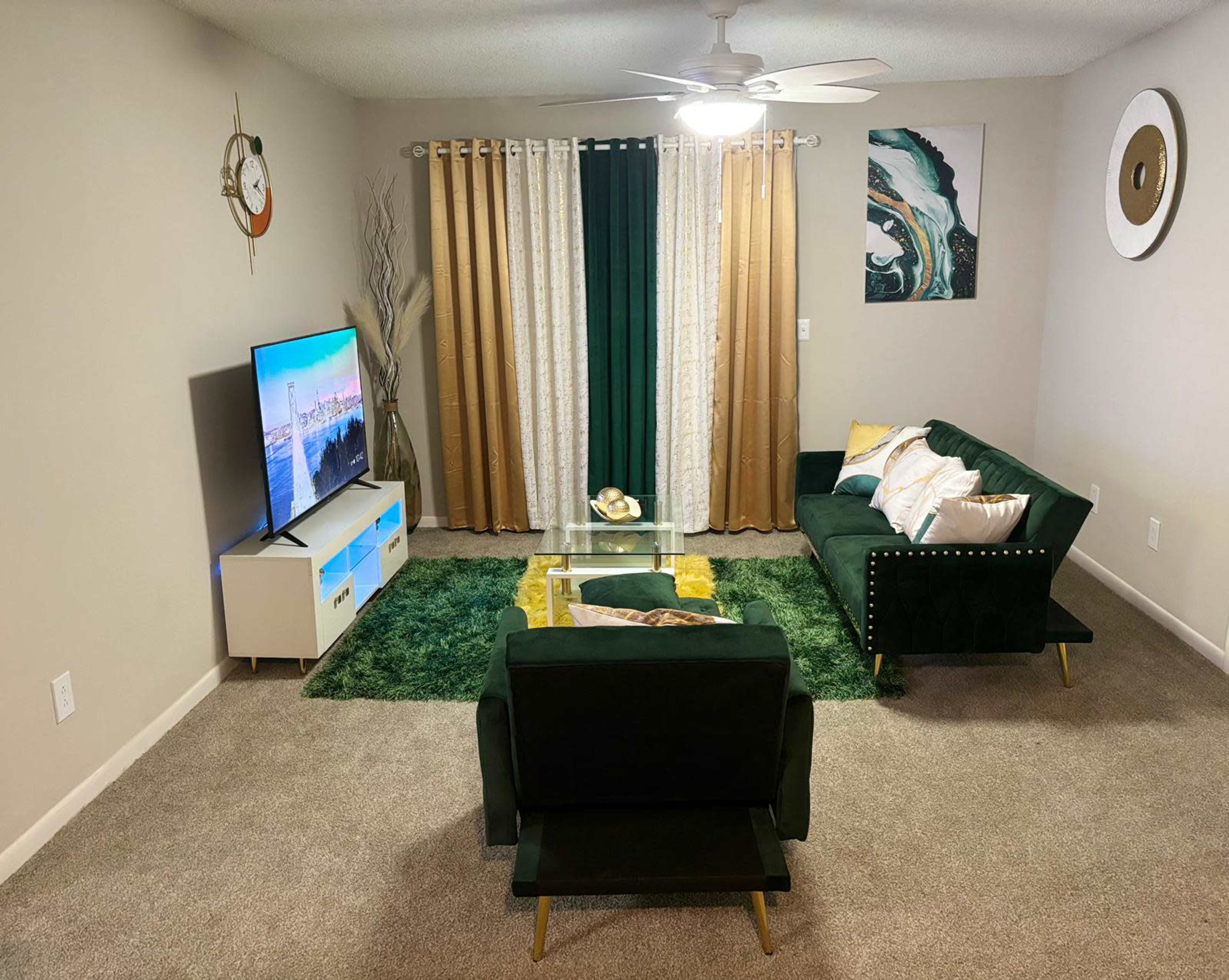 The living room features a green couch, a matching armchair, a glass coffee table, and a large television against a wall decorated with curtains and wall art.