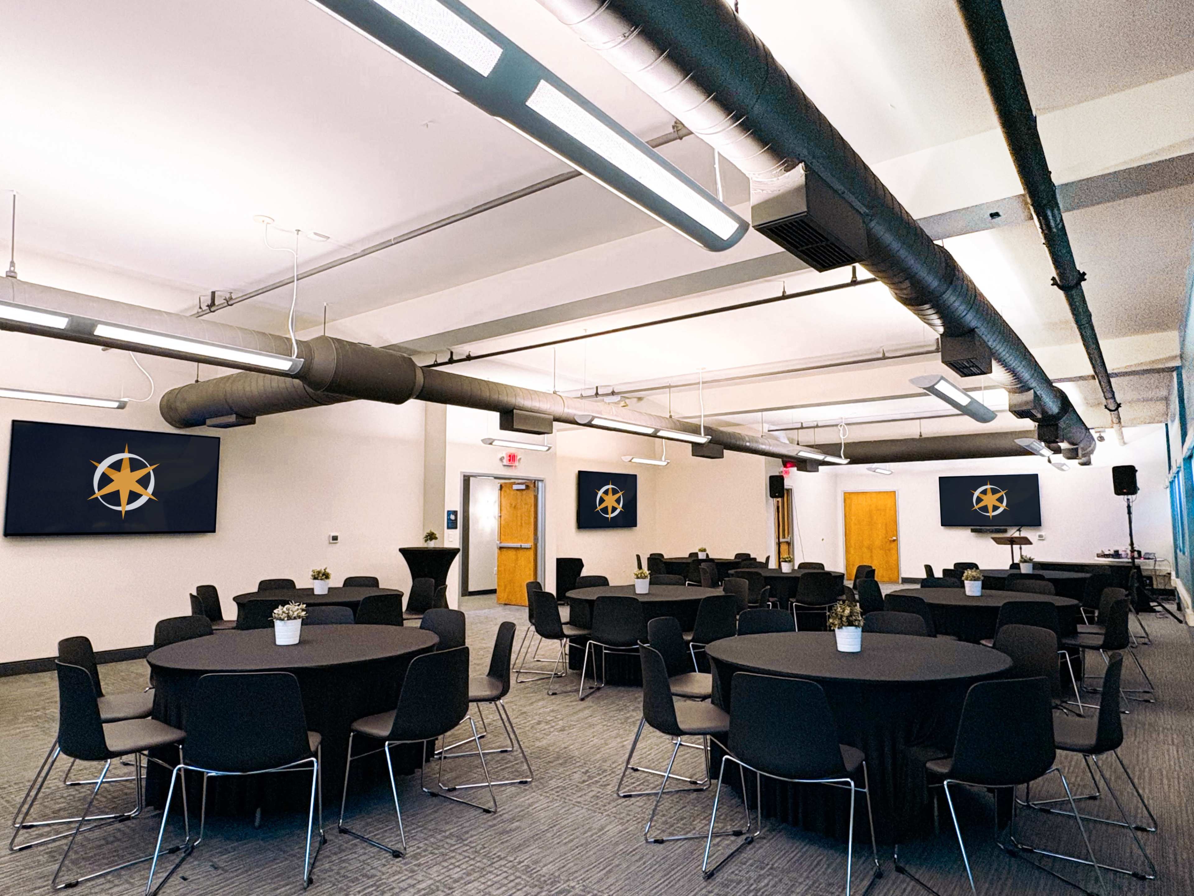 The image shows a modern conference room with several round black tables and chairs, along with large screens displaying a logo on the walls.