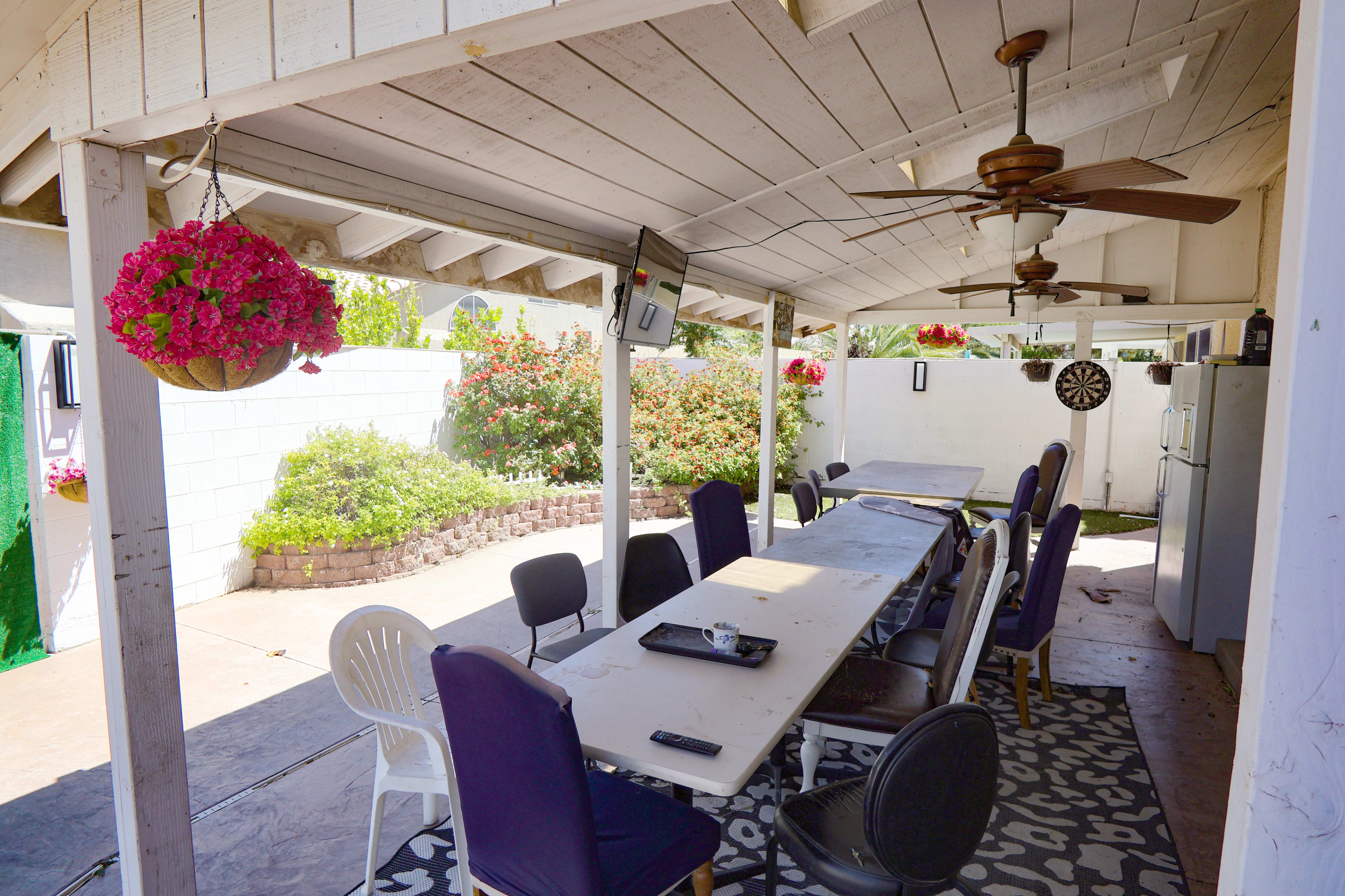 The image shows an outdoor patio area featuring a long table with several chairs, a ceiling fan, and hanging flower baskets.