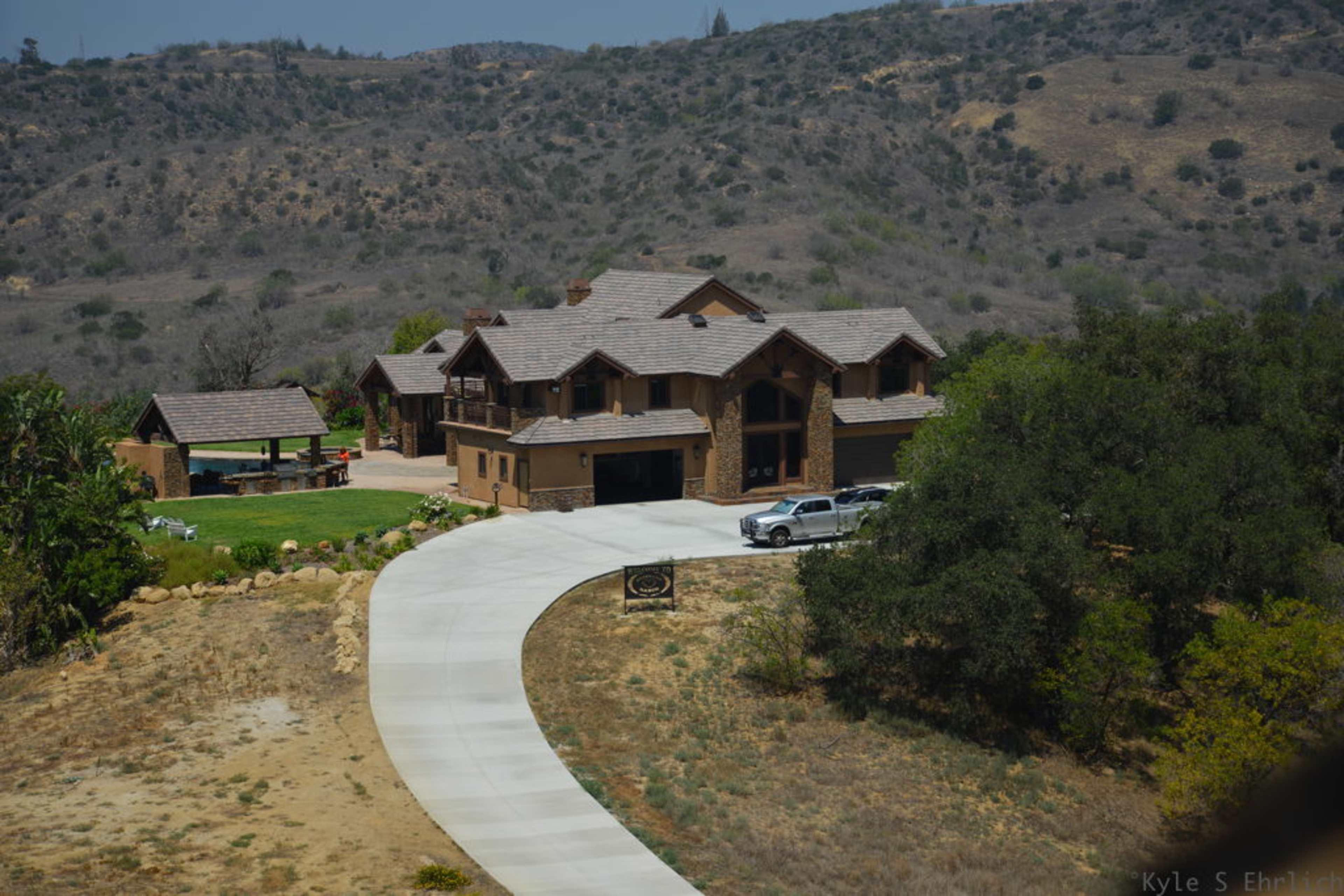The image shows a large, multi-story house with a circular driveway, surrounded by sparse landscaping and rolling hills in the background.
