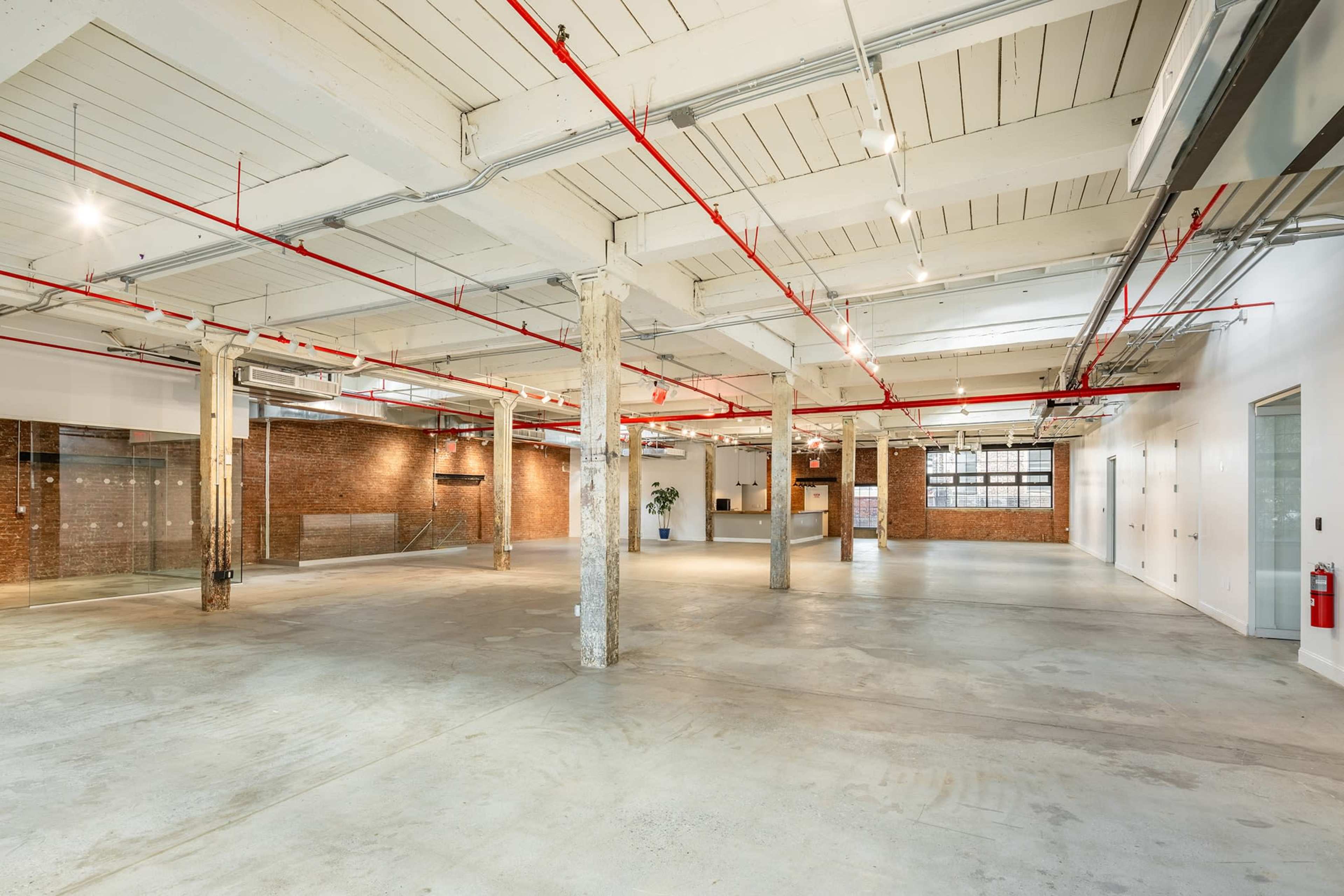 An empty industrial-style interior space with exposed concrete flooring, brick walls, and visible overhead pipes and lighting.