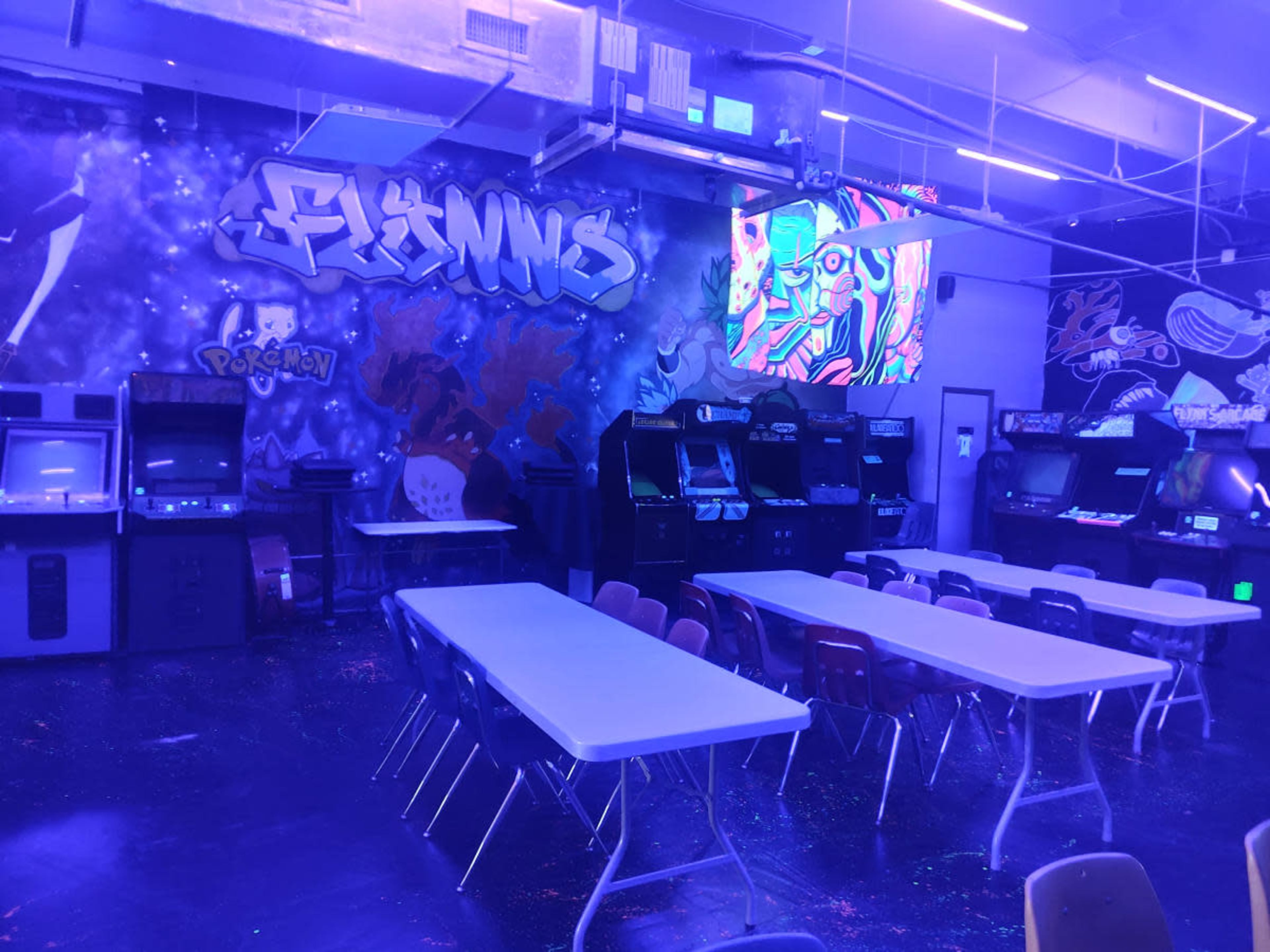 The image shows an arcade room with rows of gaming machines, tables, and colorful wall murals.