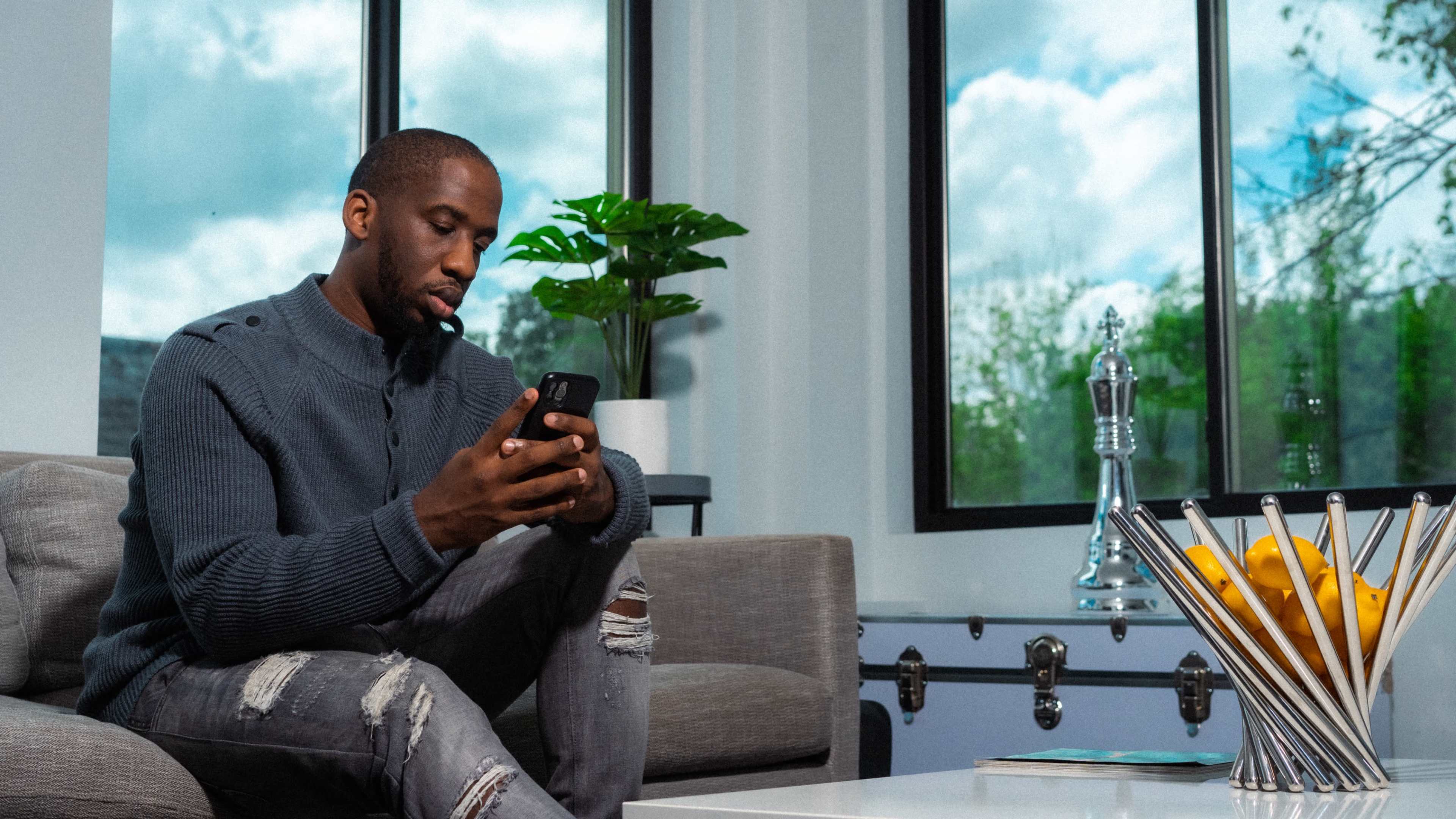 A man sits on a sofa, focused on his phone, with a plant and a bowl of lemons on a coffee table nearby.