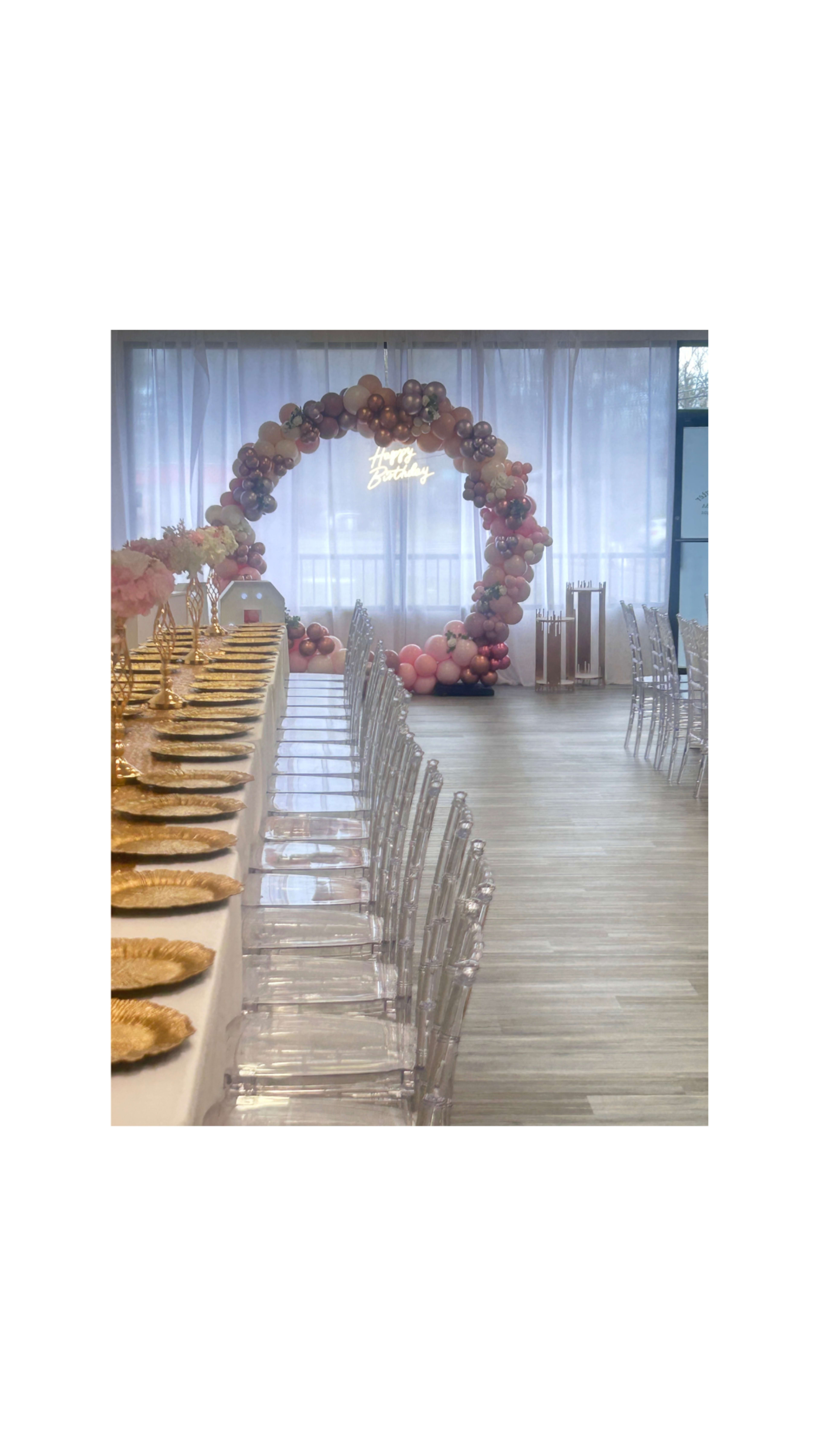 A long table set with gold plates and clear chairs is arranged in a brightly lit room featuring a colorful balloon arch with a neon "Happy Birthday" sign in the background.