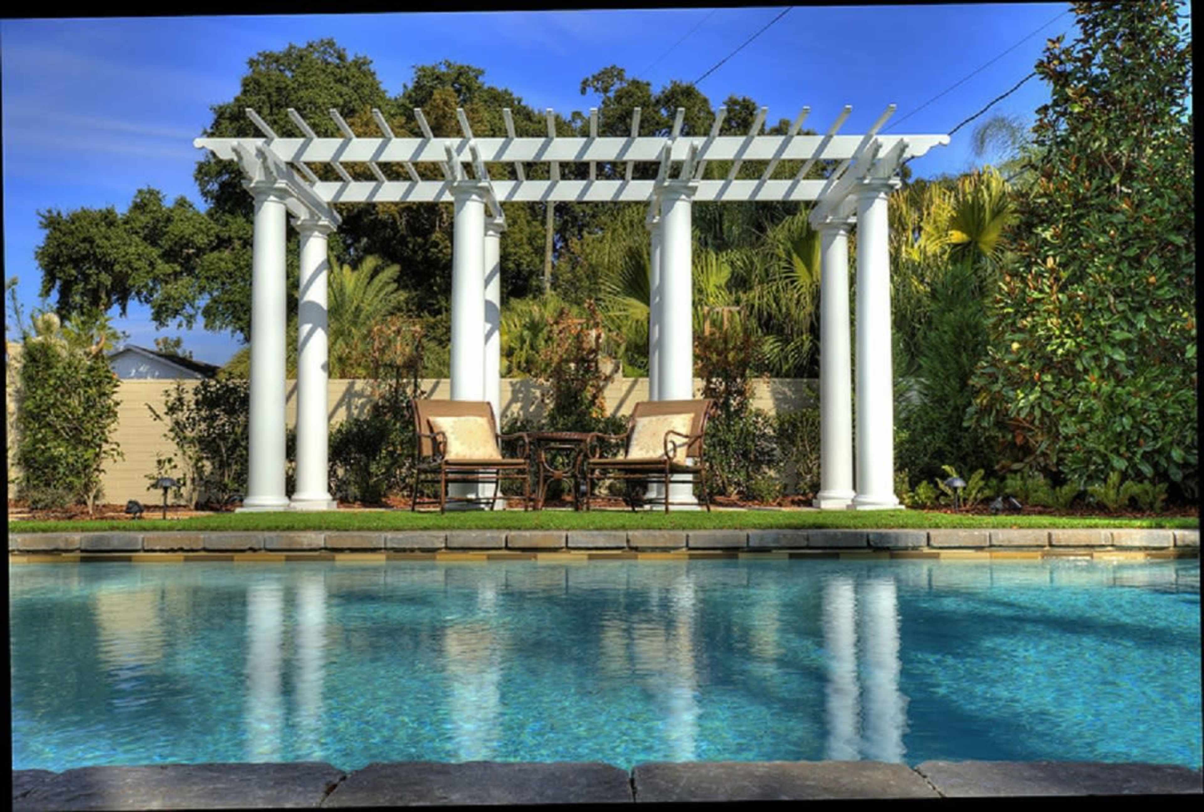 A tranquil swimming pool with a pergola supported by white columns in the background, surrounded by lush greenery.