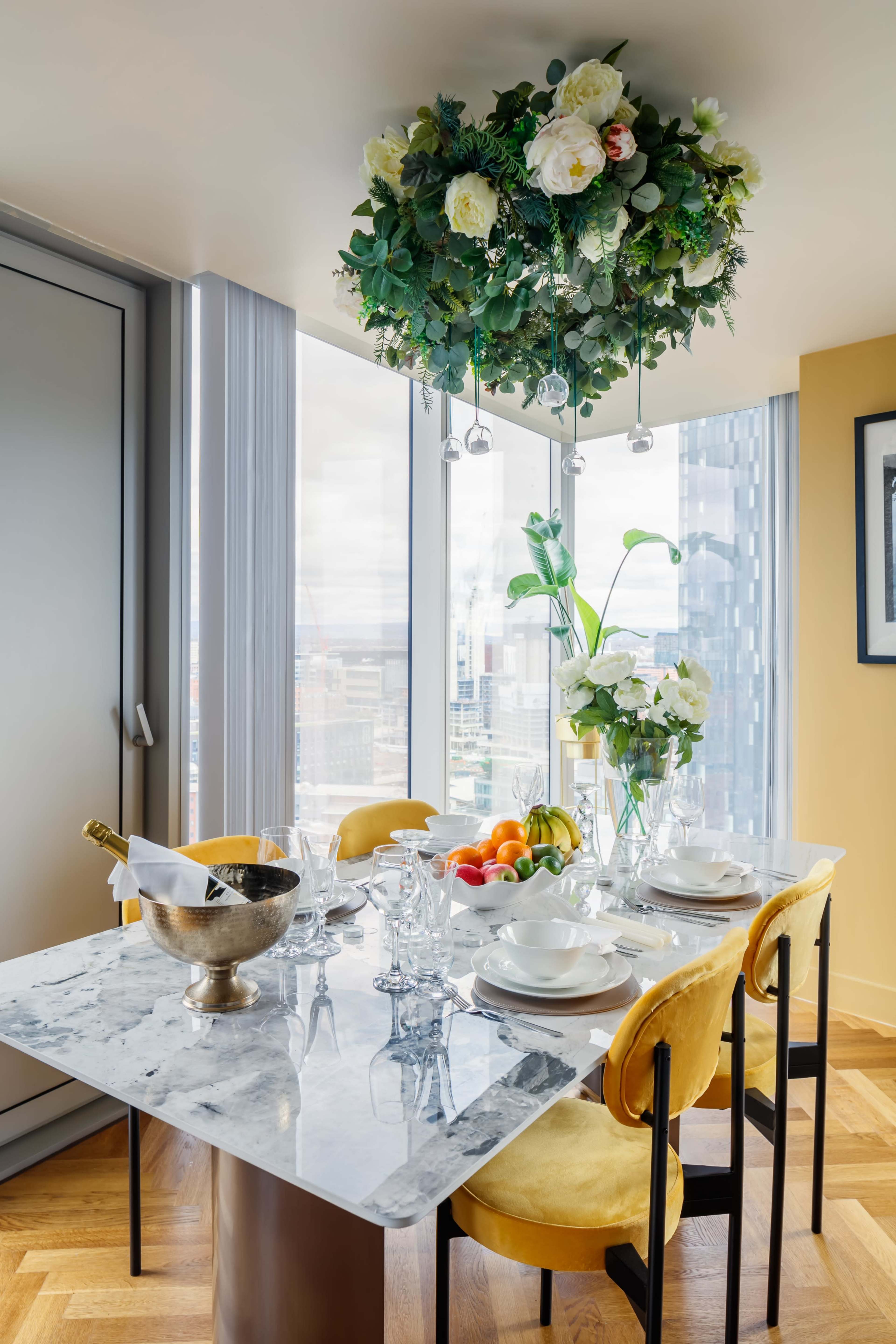 A modern dining table is set with plates and glassware, complemented by a floral chandelier and a bouquet, with a view of the city skyline visible through large windows.