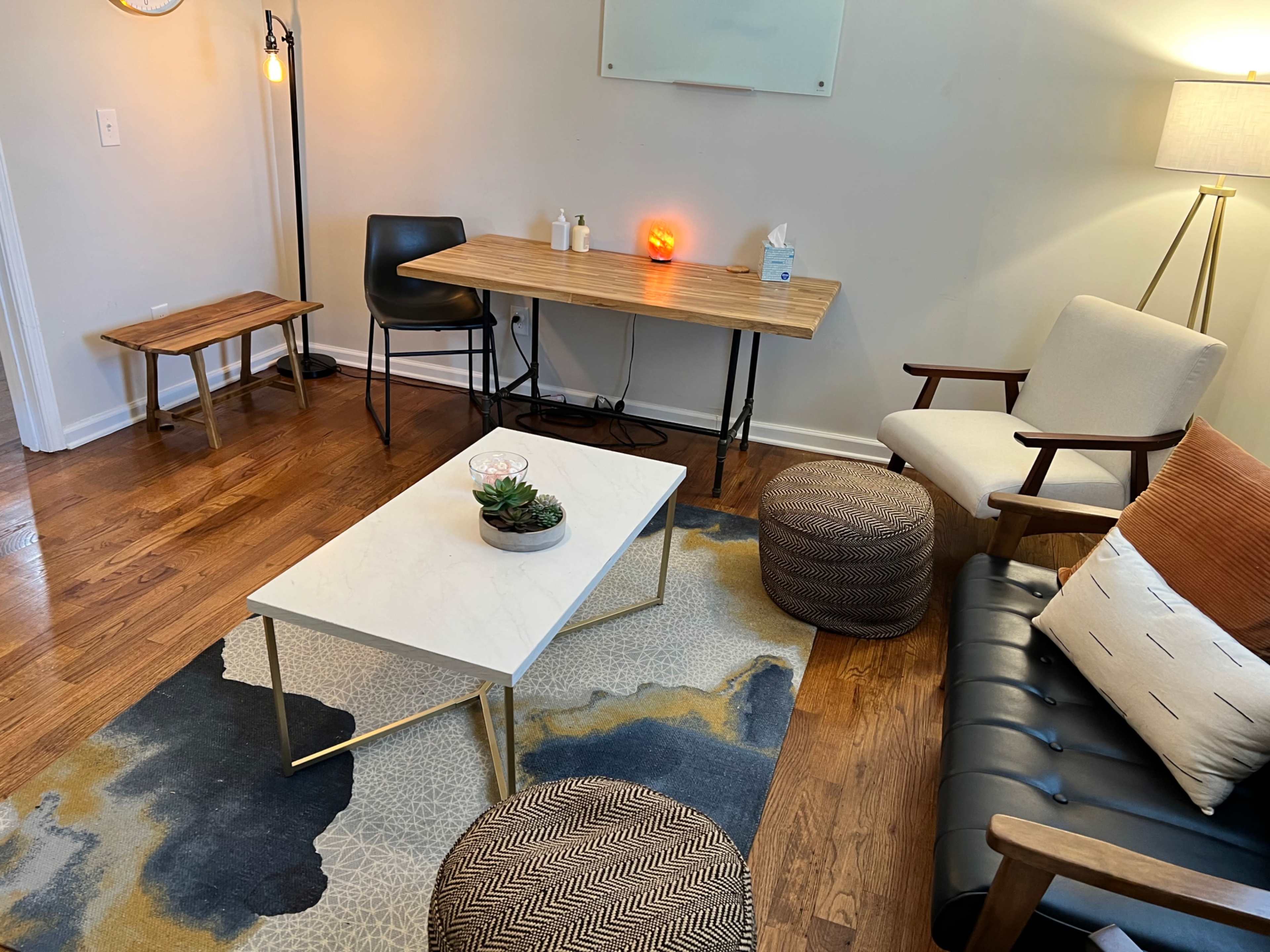 The image shows a cozy living space featuring a wooden table and a black chair, a white coffee table, a patterned rug, and various seating options including an armchair and poufs.