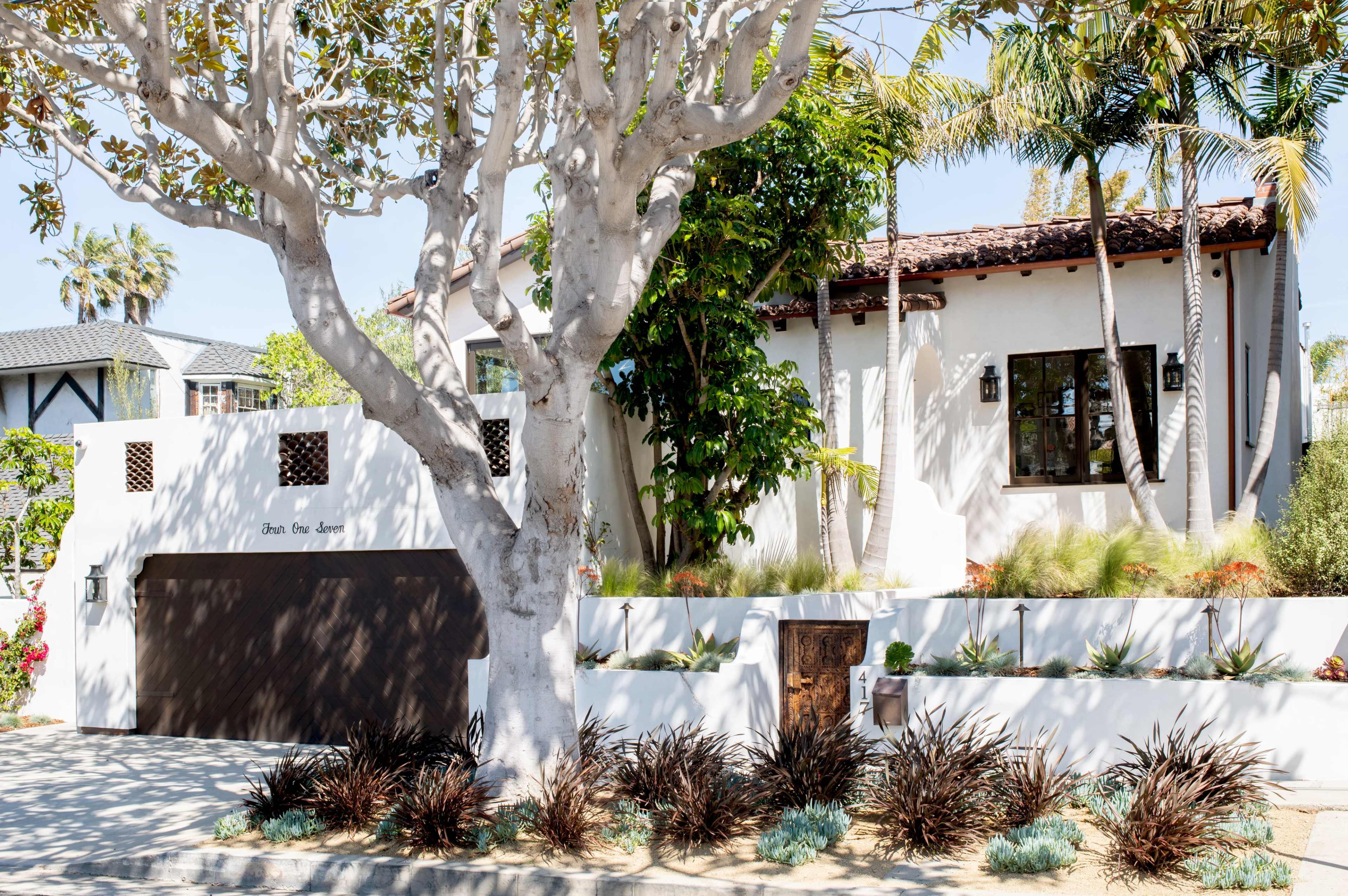 A white house with a dark garage door, surrounded by palm trees and landscaping, sits behind a low wall.