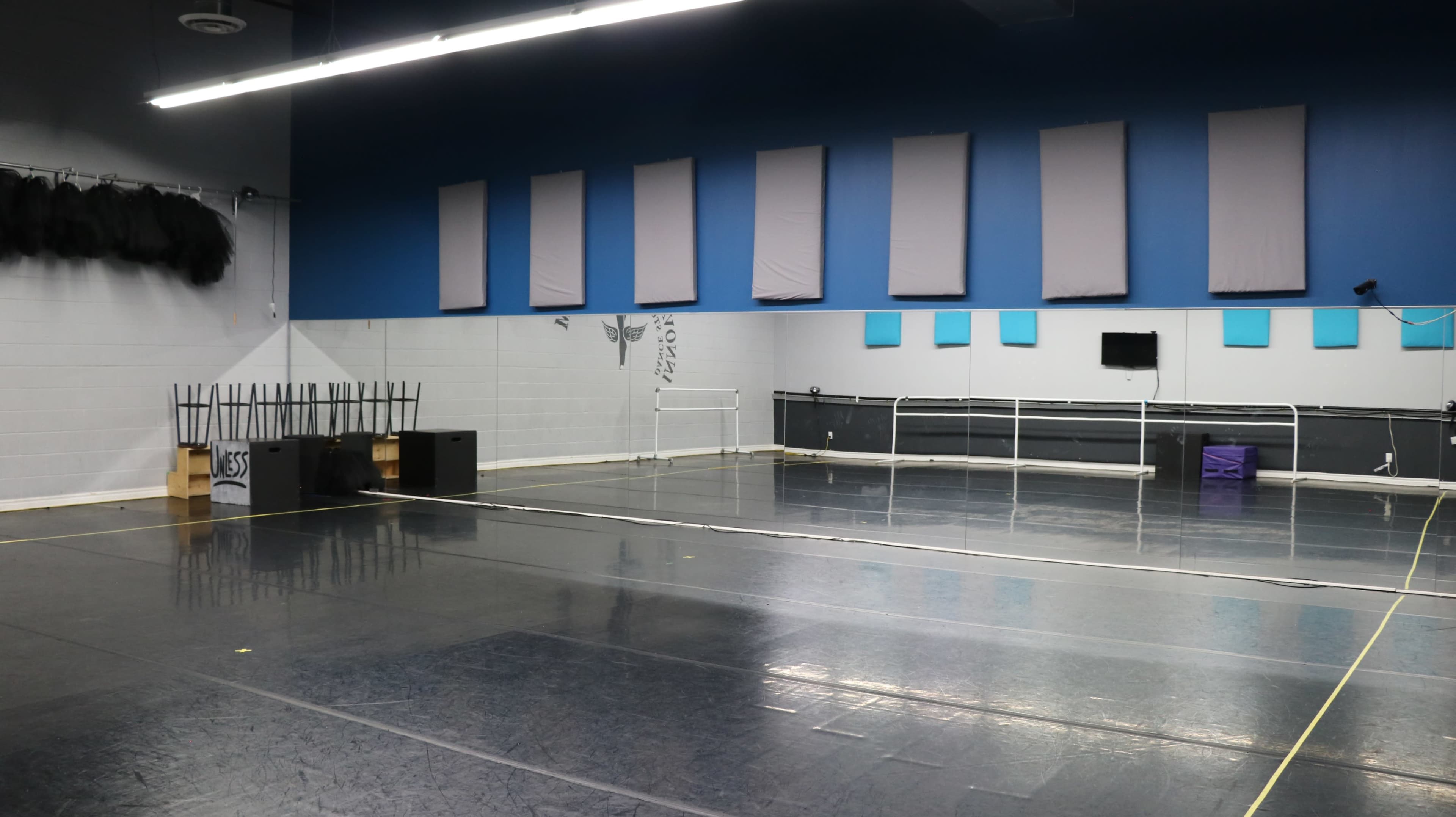 The image shows a large, empty dance studio with a polished floor, mirrors along one wall, and a blue accent wall featuring square panels.