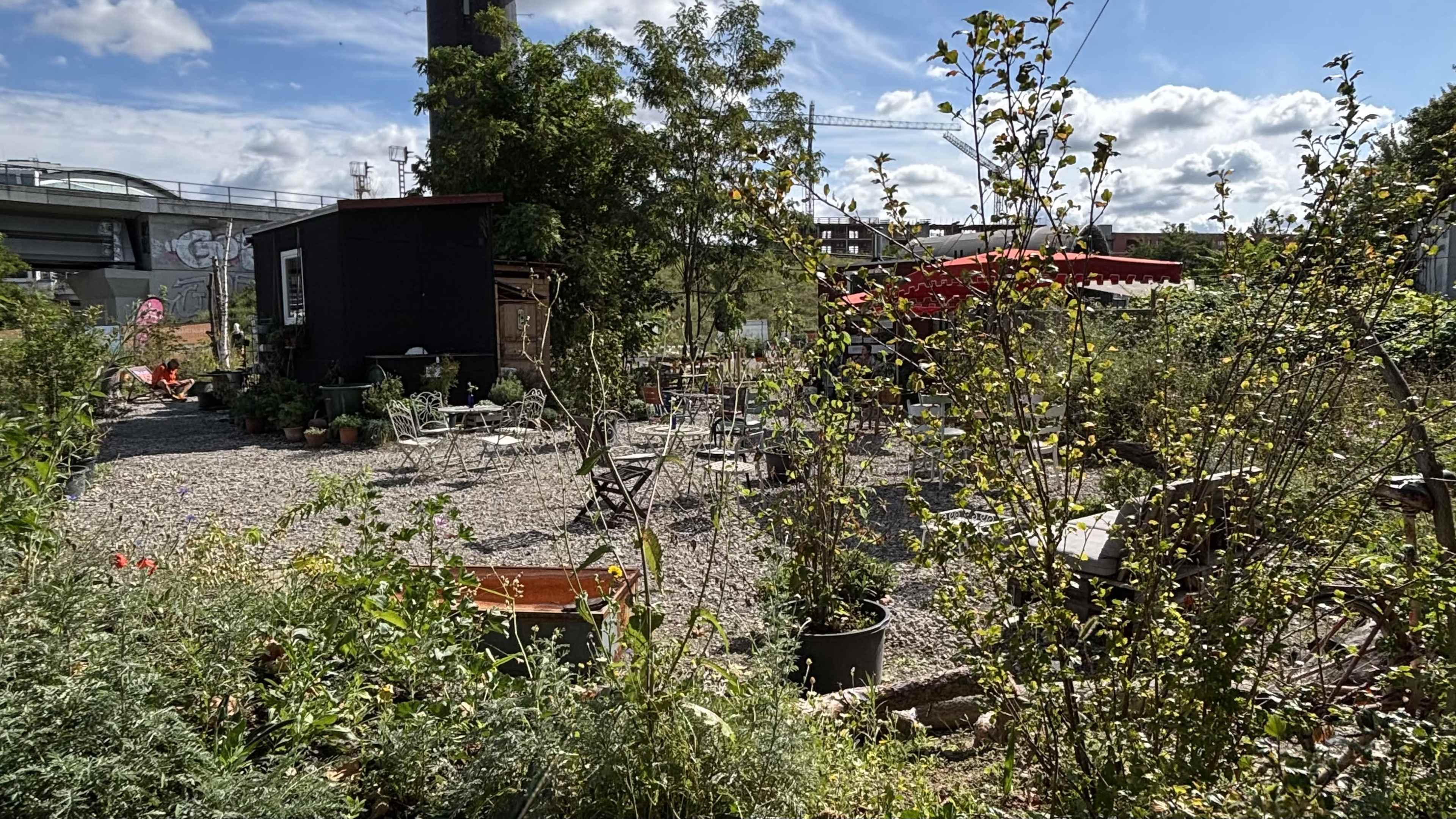The scene features a garden area filled with various plants and furniture set on gravel, with a black building in the background and a red canopy further back.