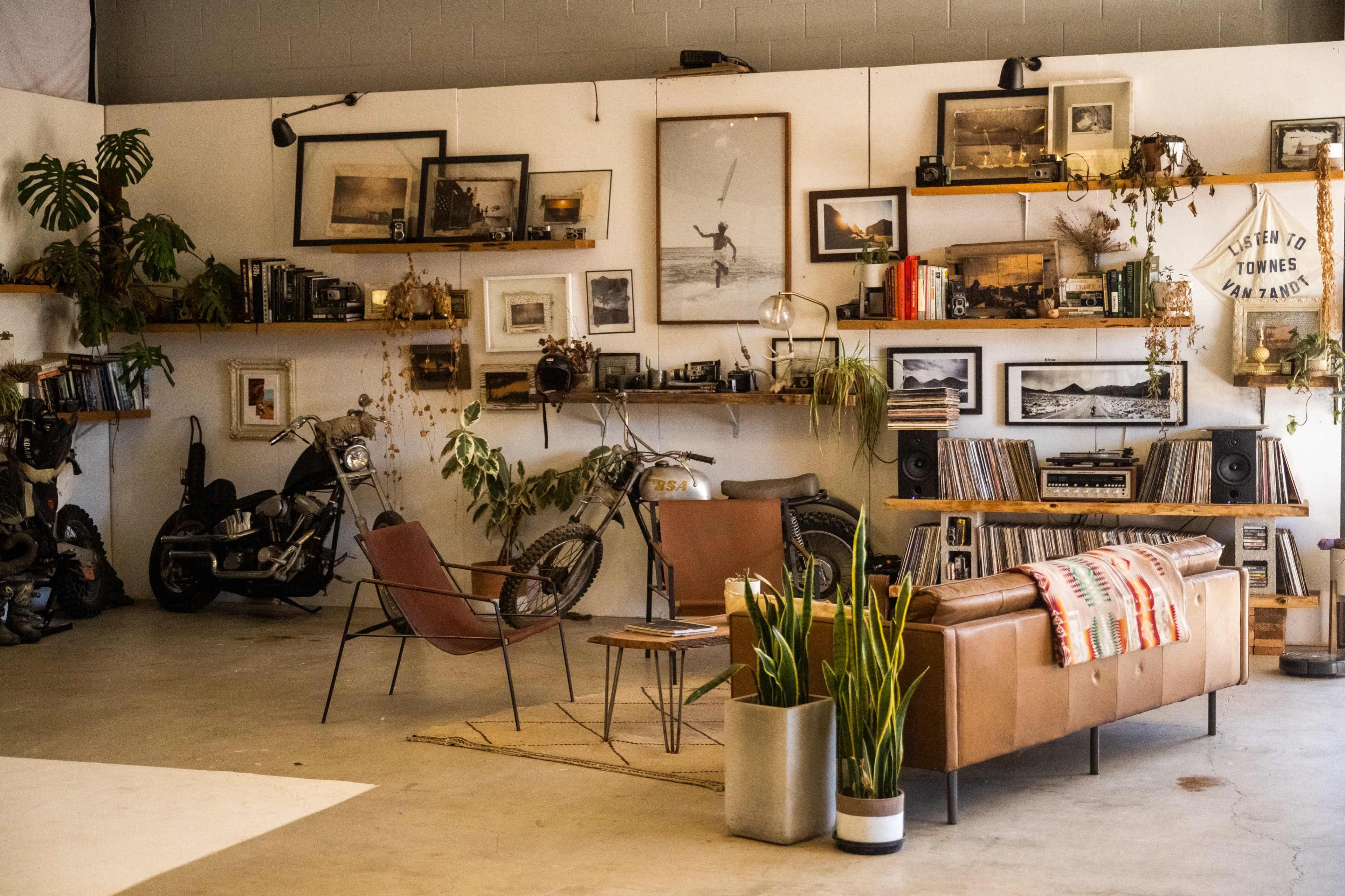 The image shows a spacious garage featuring vintage motorcycles, a cozy seating area with a leather sofa and armchair, and a wall adorned with framed photographs and shelves of records.