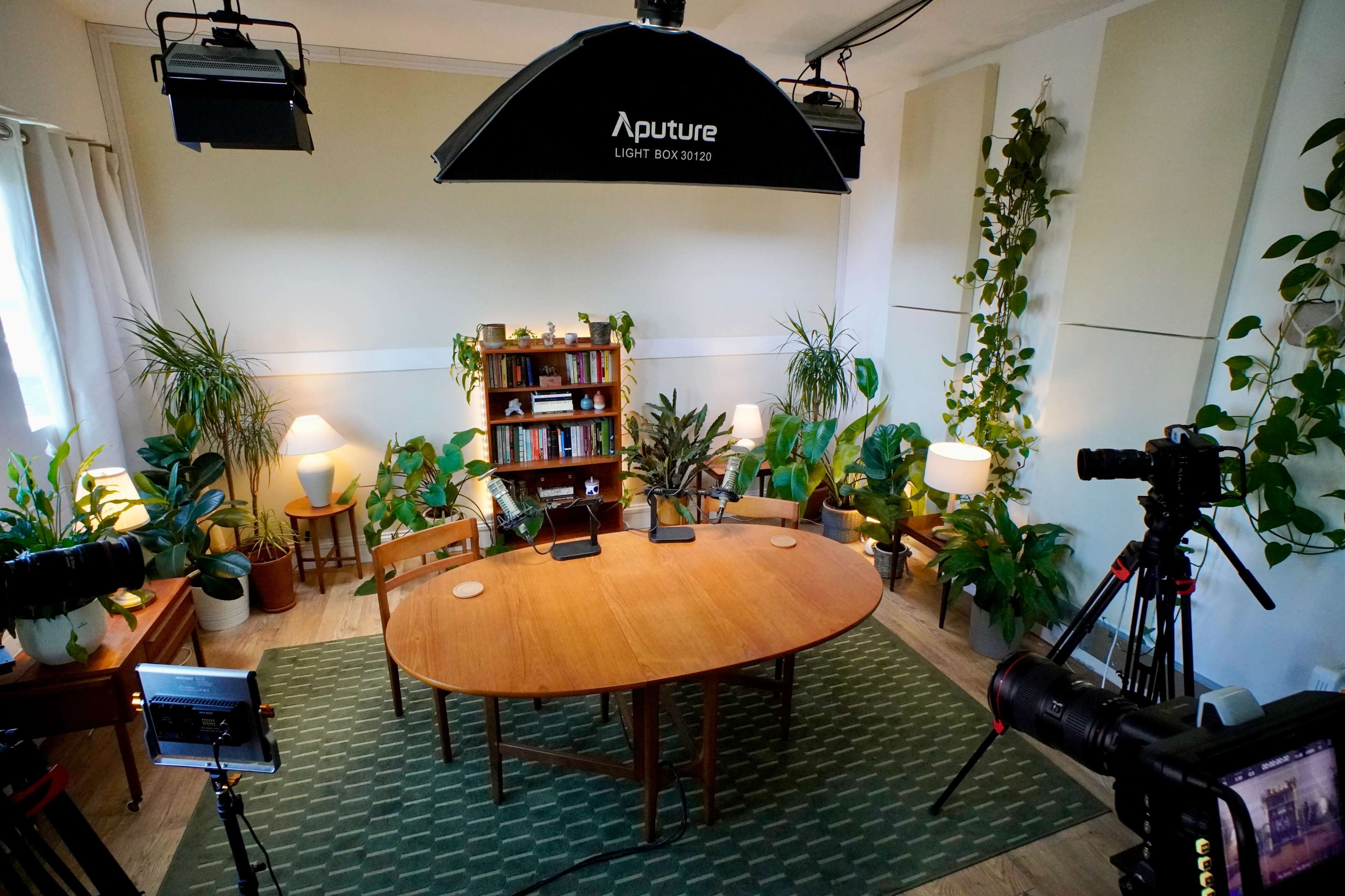 The image shows a well-lit studio setup for a discussion or interview, featuring a wooden table surrounded by chairs, plants, and cameras.