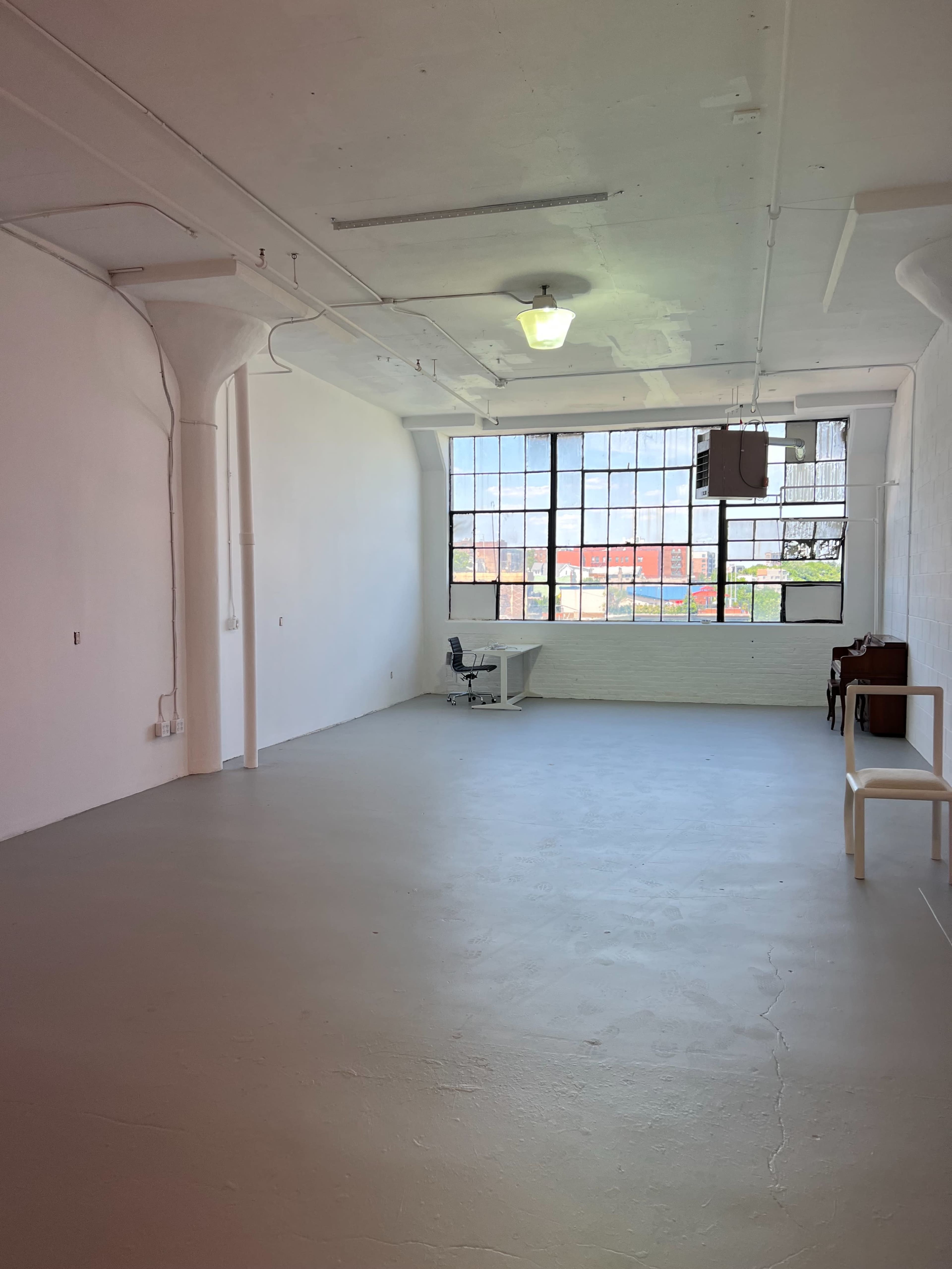 A spacious, empty room with a large window and minimal furnishings, featuring a desk and a chair.