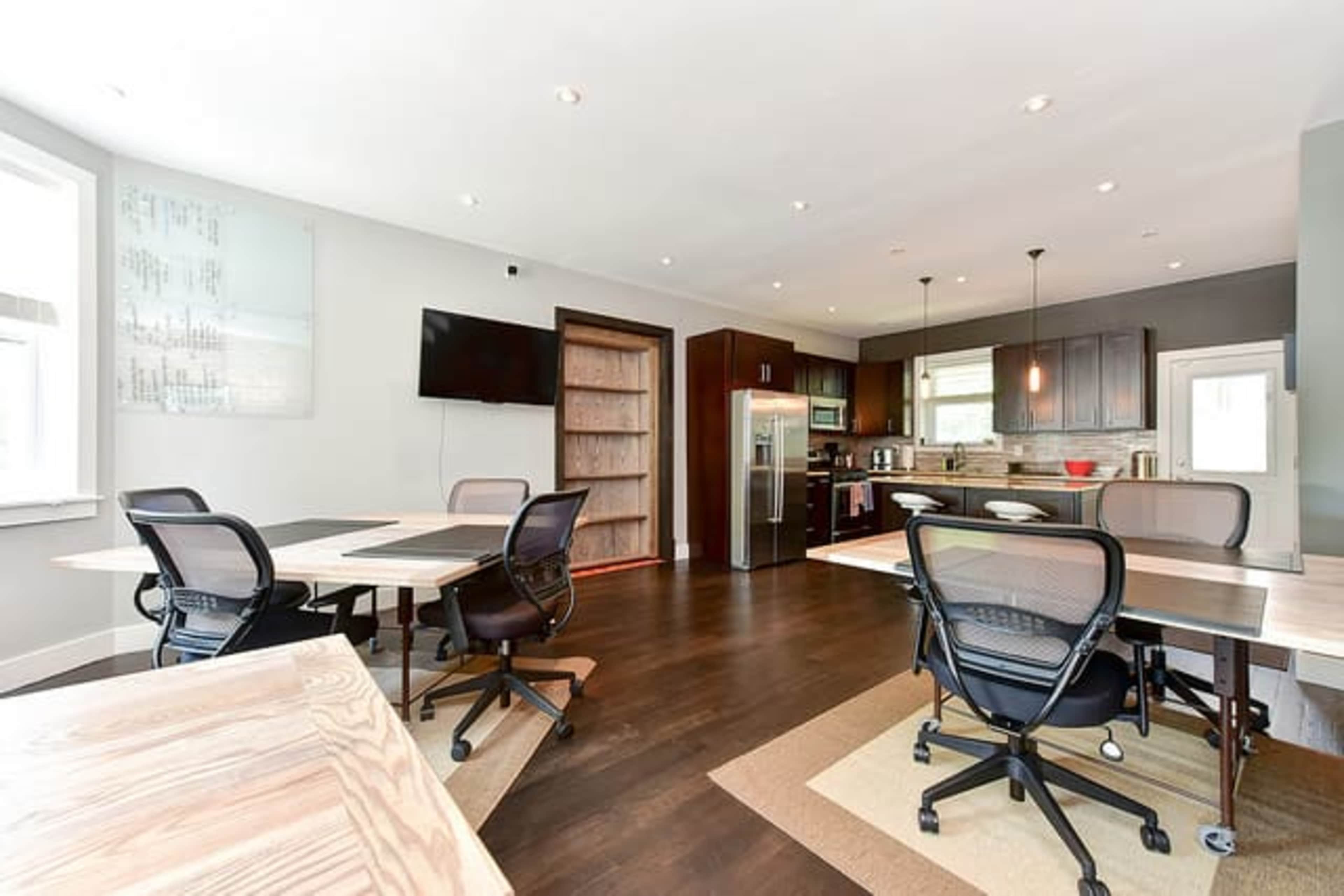 A modern office space with several work tables and chairs, a kitchenette area, and a large television mounted on the wall.