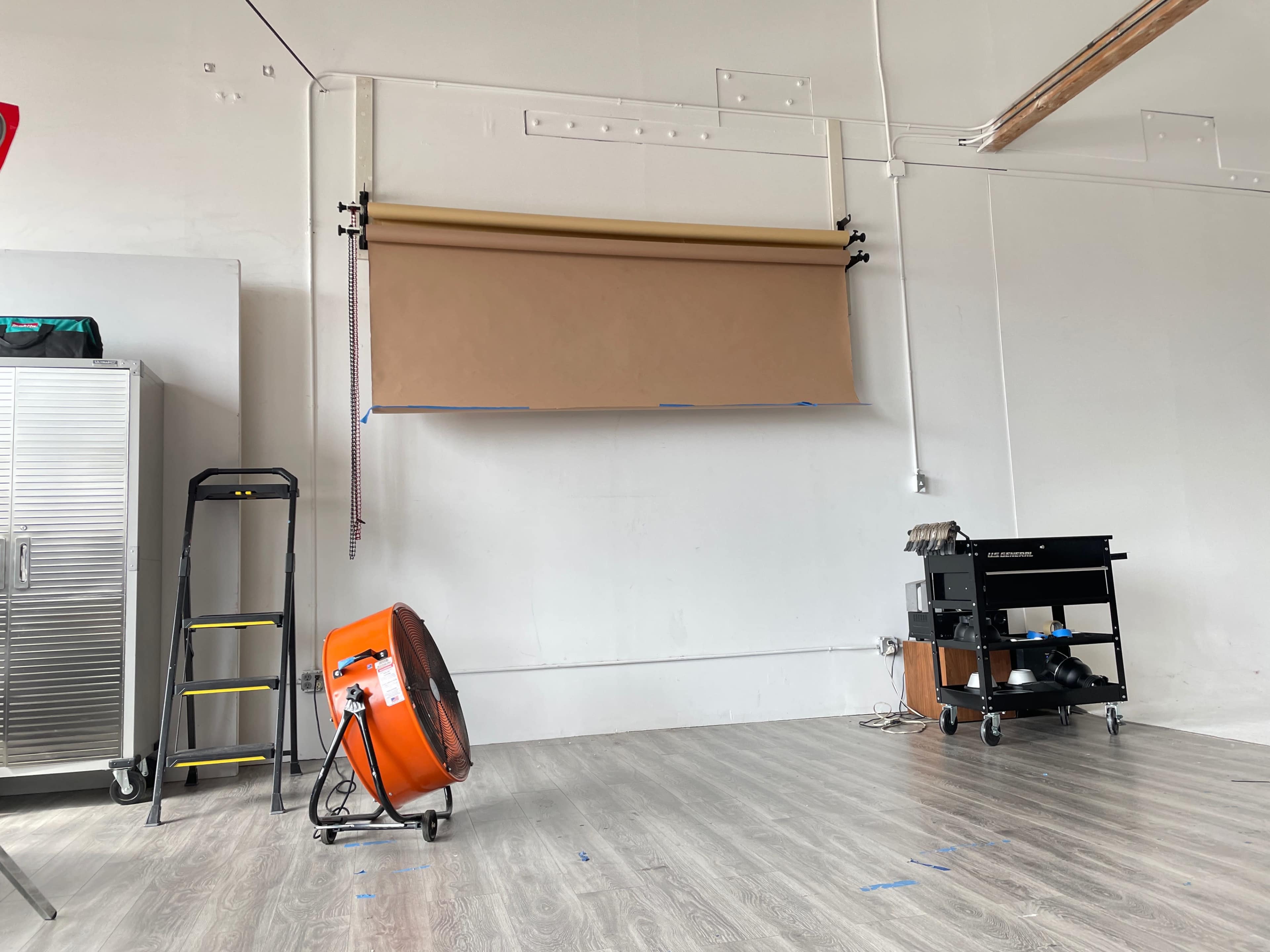 A studio space with a large brown paper backdrop mounted on the wall, a yellow ladder, an orange fan, and a black equipment cart.