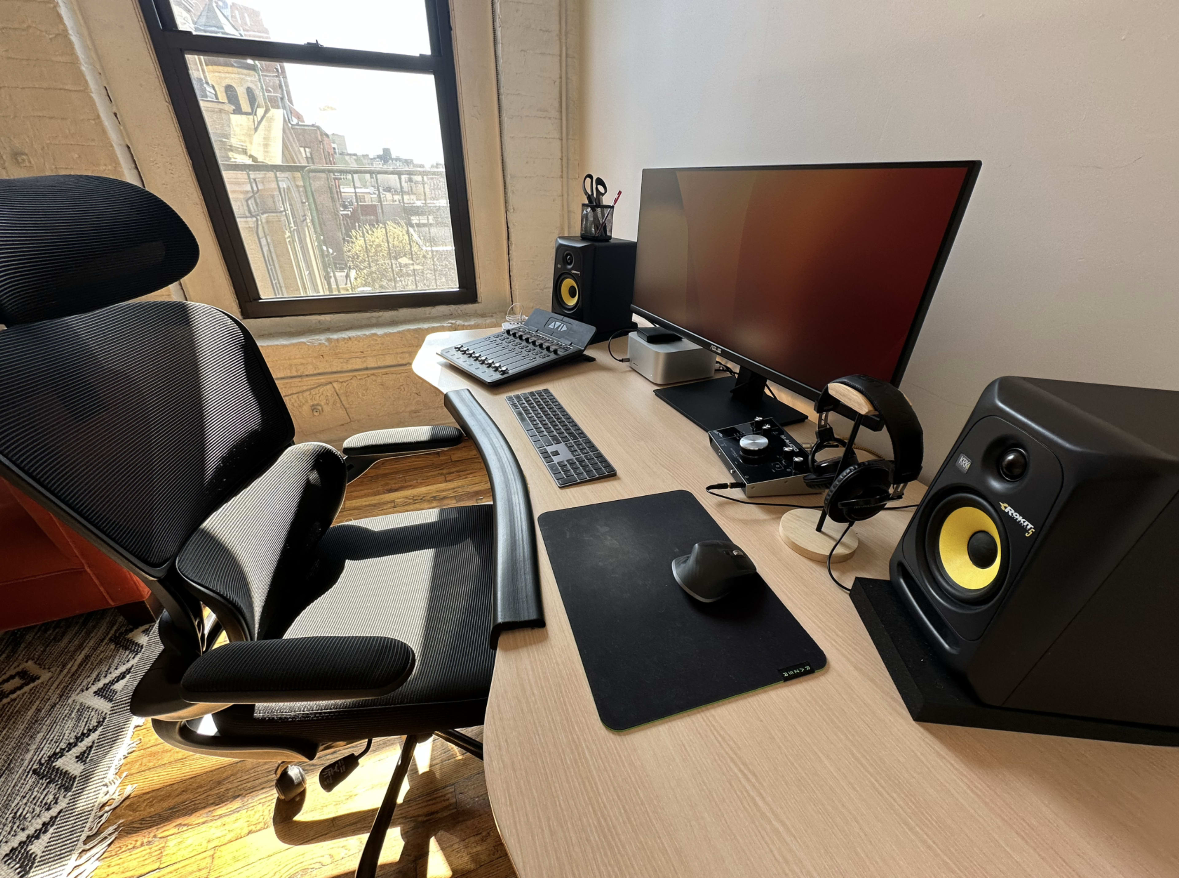 The image shows a modern workspace with an ergonomic chair, a large monitor, a keyboard, a mouse, and two studio monitors on a desk near a window.