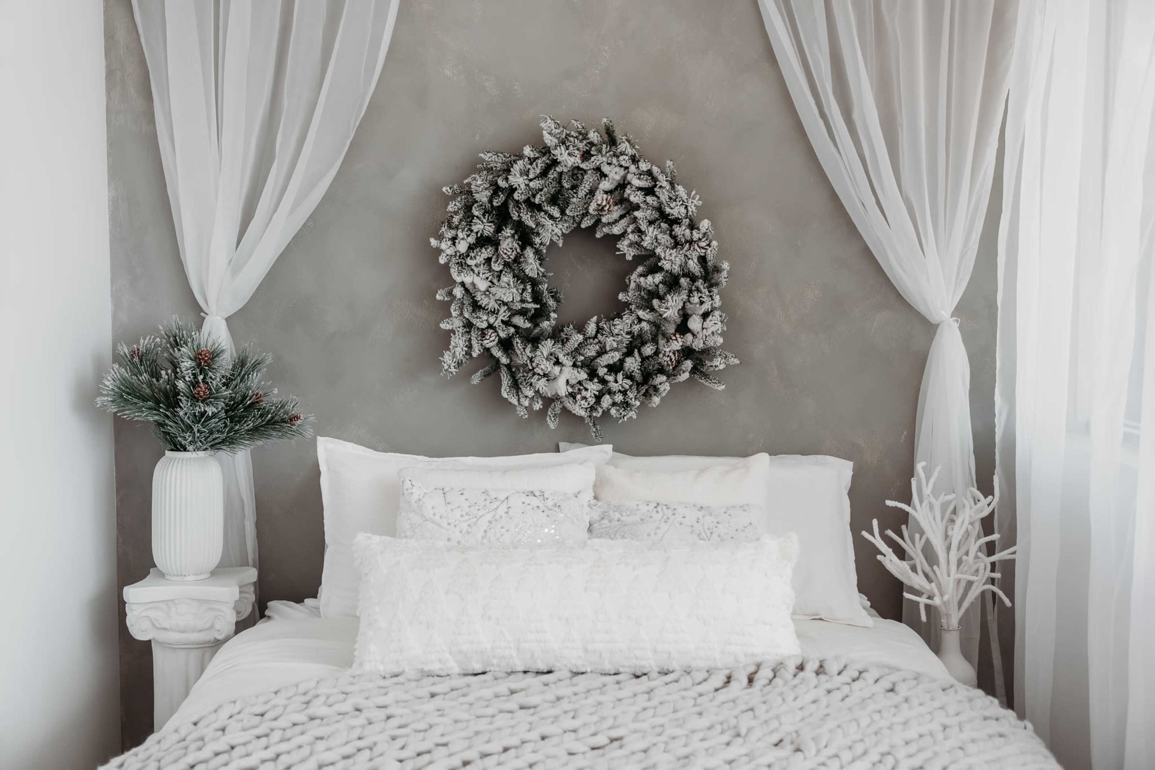 The image shows a cozy bedroom with a bed adorned with white pillows and a textured blanket, featuring a frosted wreath on the wall and decorative plants in white vases.