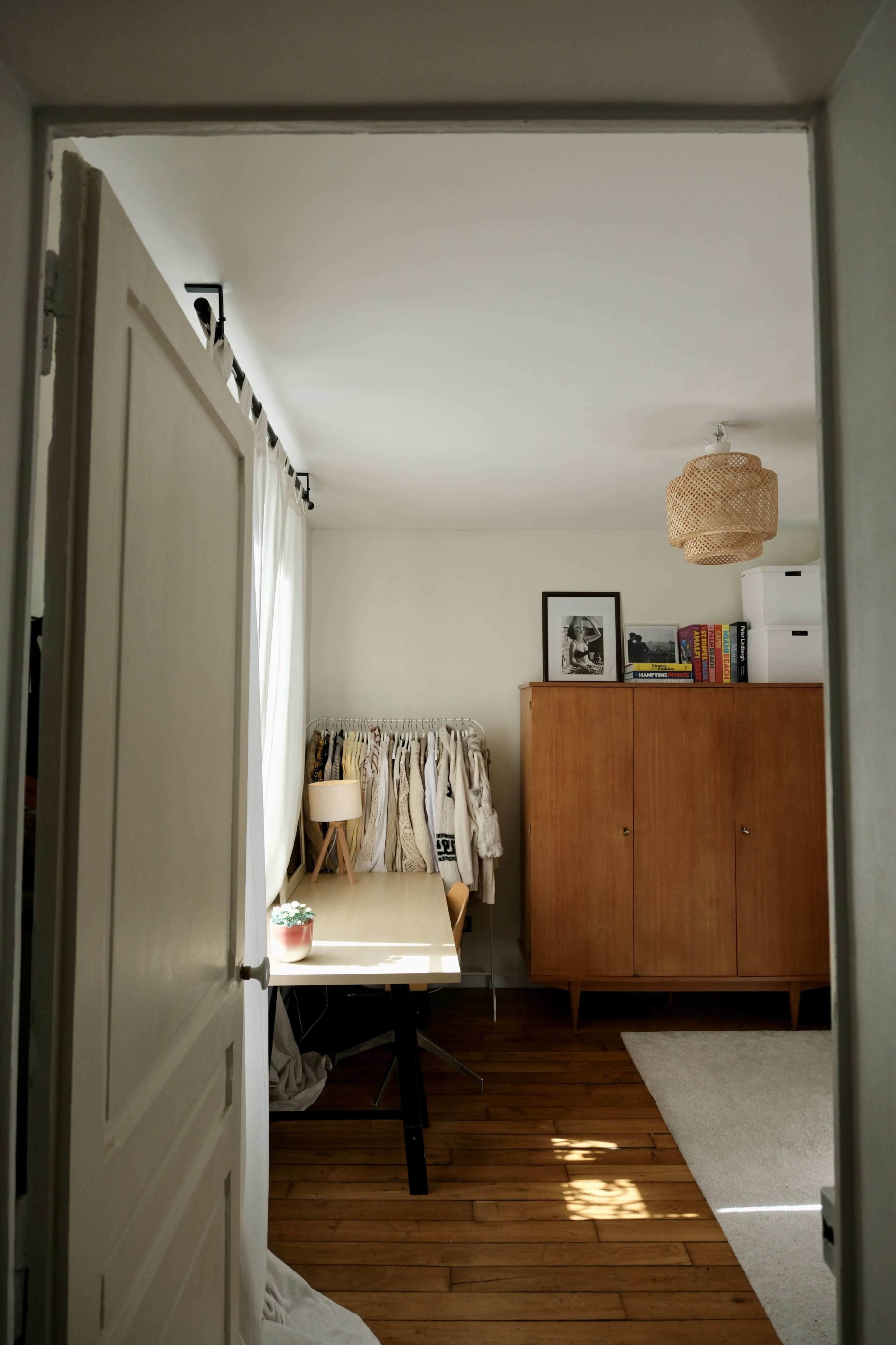 The image shows a well-lit room with a wooden floor, featuring a table, a wardrobe, and a clothing rack in the corner.
