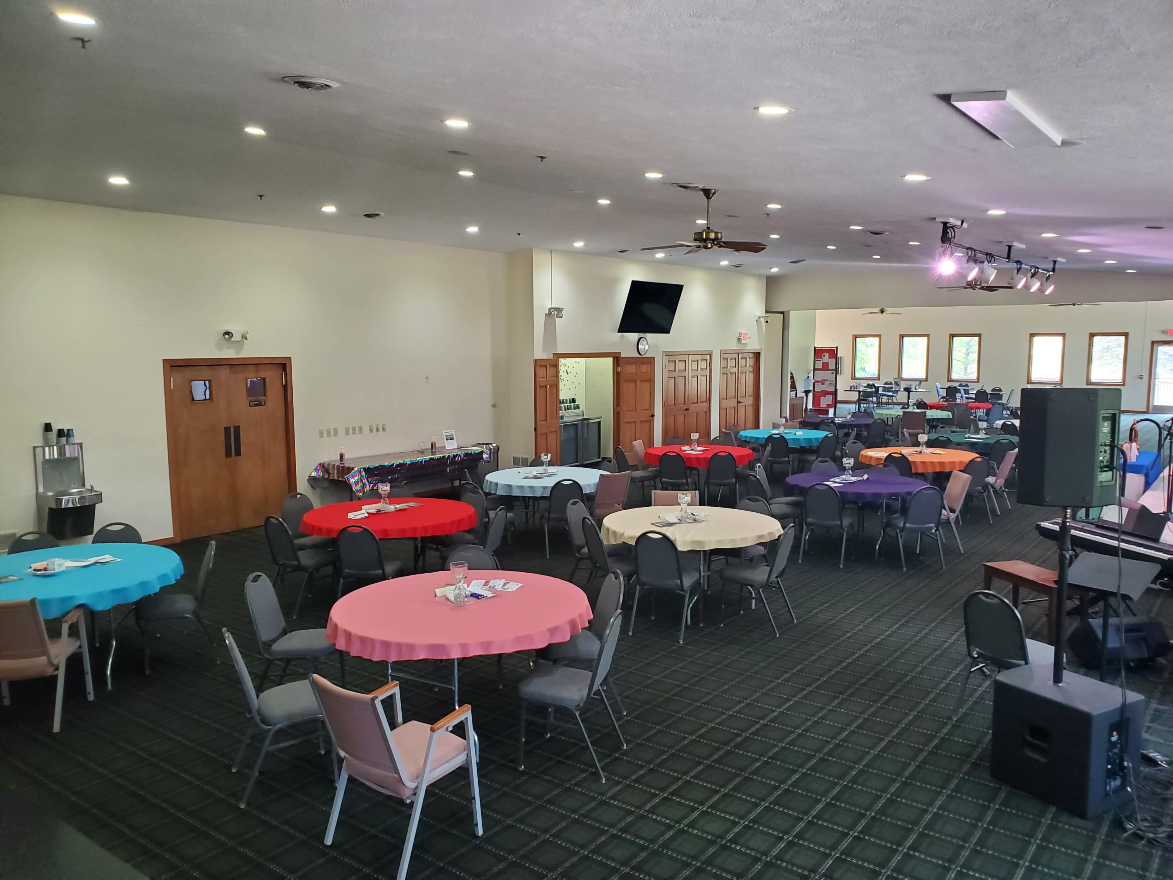 An event space is set up with multiple tables covered in colorful tablecloths, arranged around a stage area.