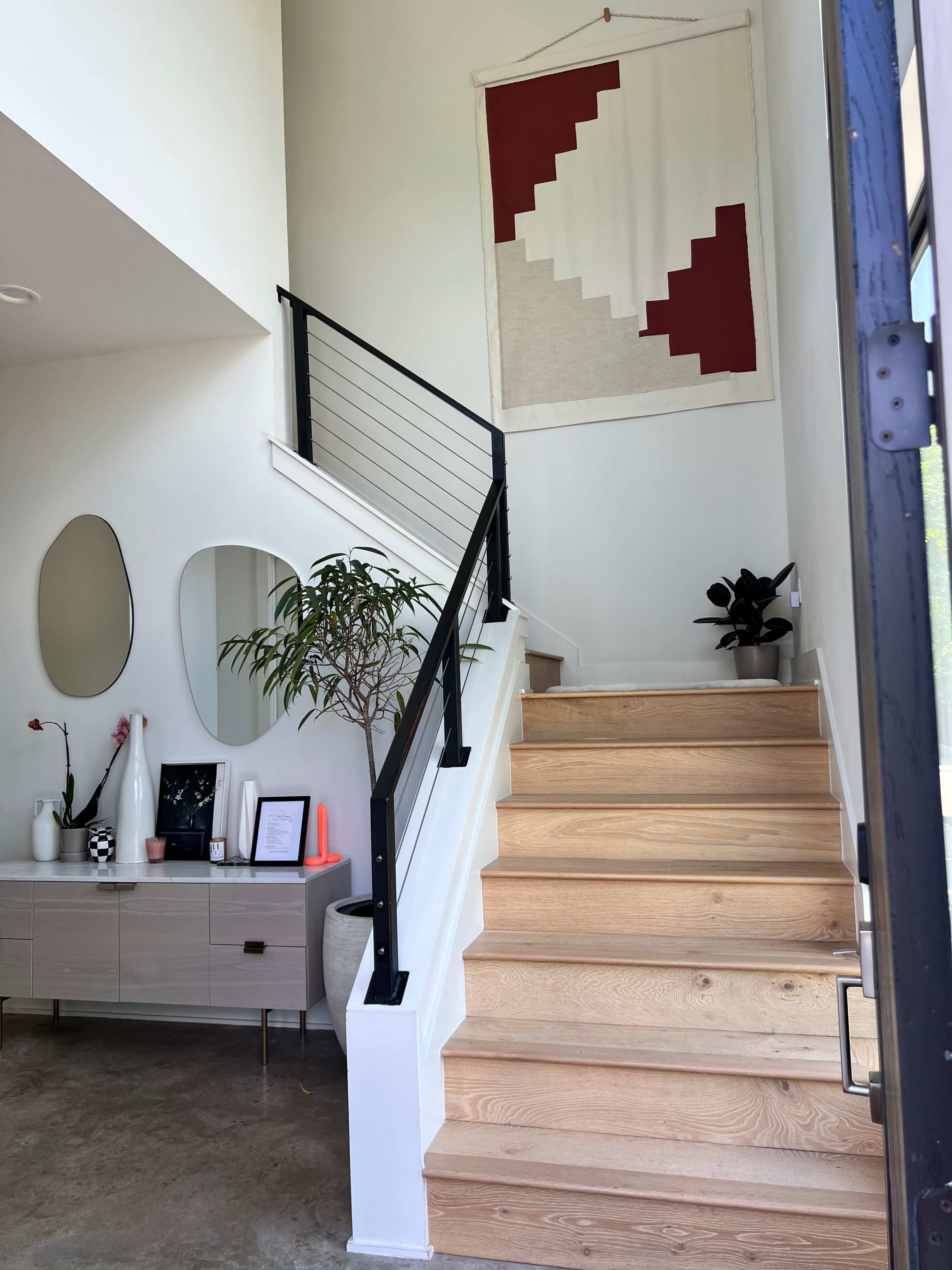 A well-lit entryway features a wooden staircase with a minimalist railing, a decorative wall hanging, and a console table with various decor items.