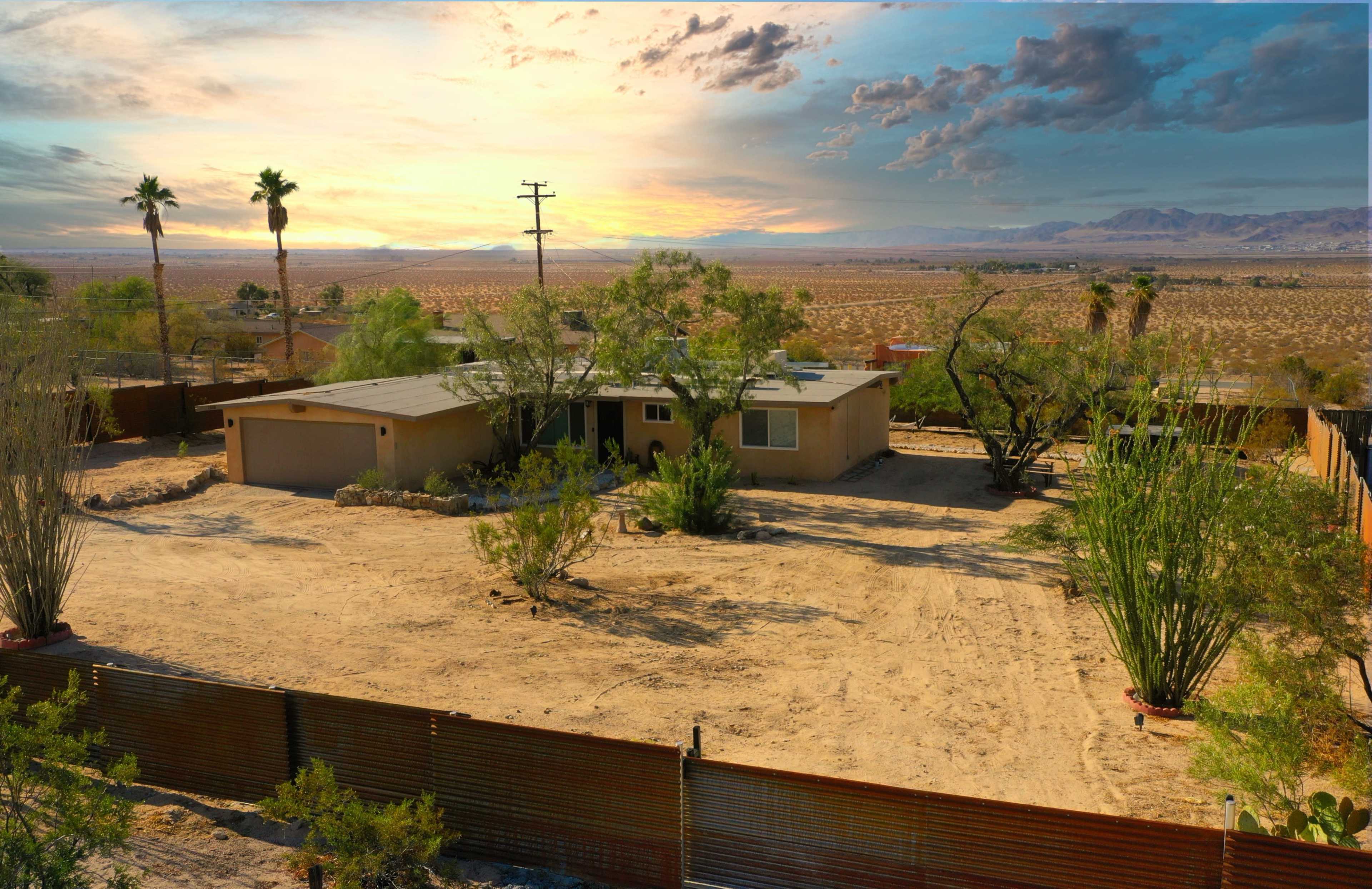 A single-story house with a flat roof is surrounded by sparse vegetation and fencing, set against a backdrop of mountains and a sunset sky.