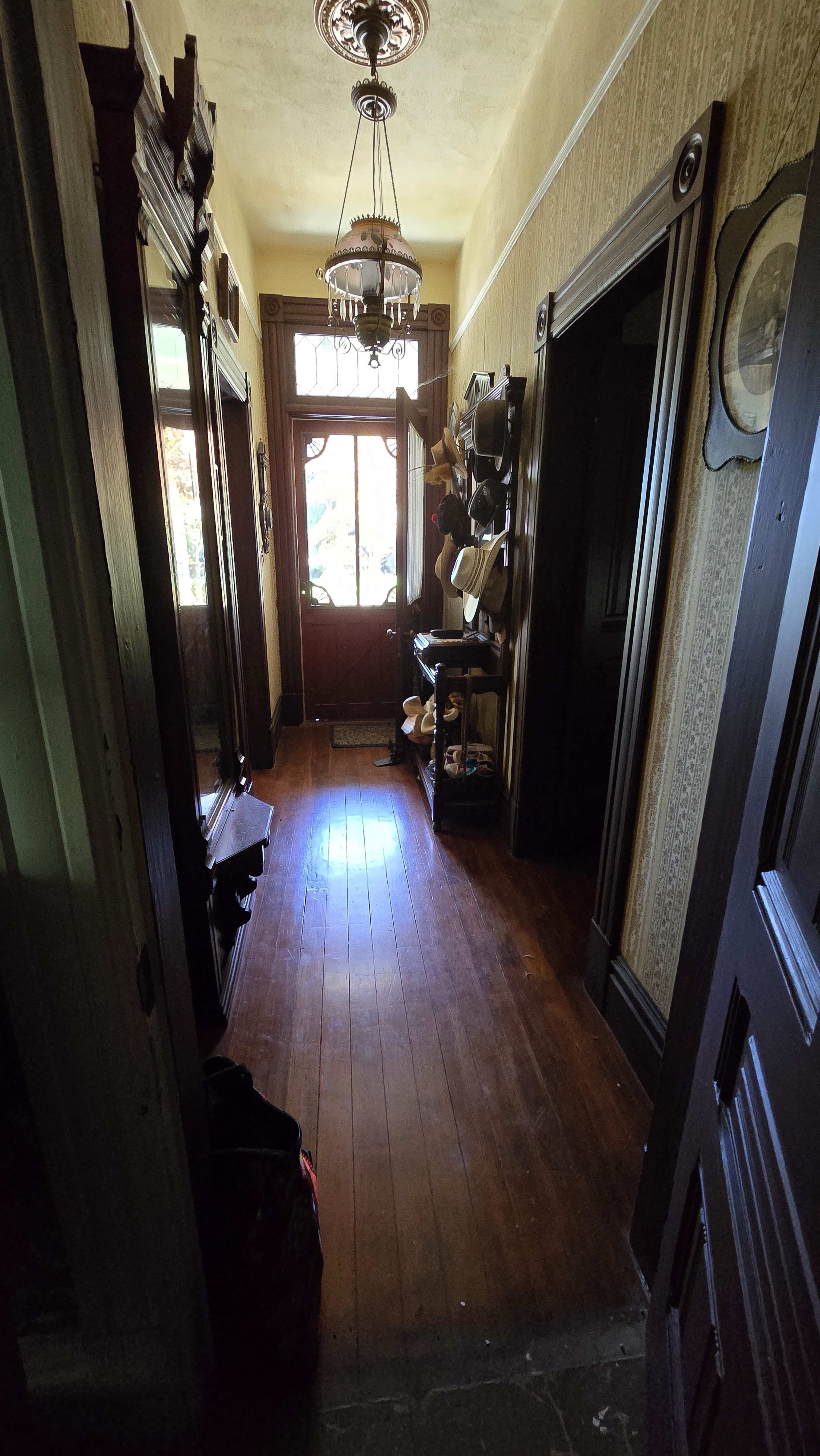 A narrow hallway with wooden flooring, framed by ornate wooden doorways and illuminated by light coming in from a glass-paneled front door.