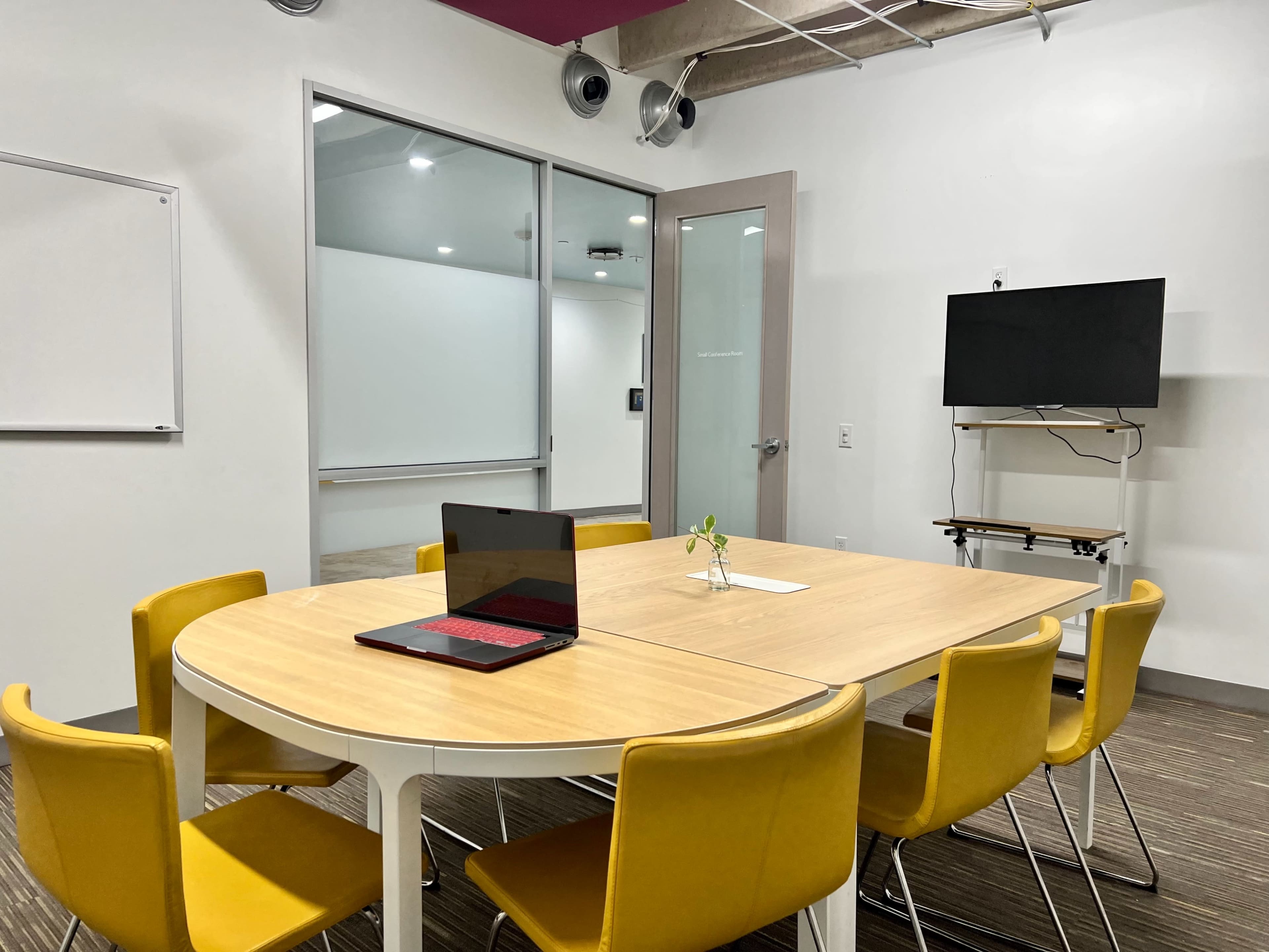 A conference room features a round table surrounded by yellow chairs, with a laptop open on the table and a television mounted on the wall.