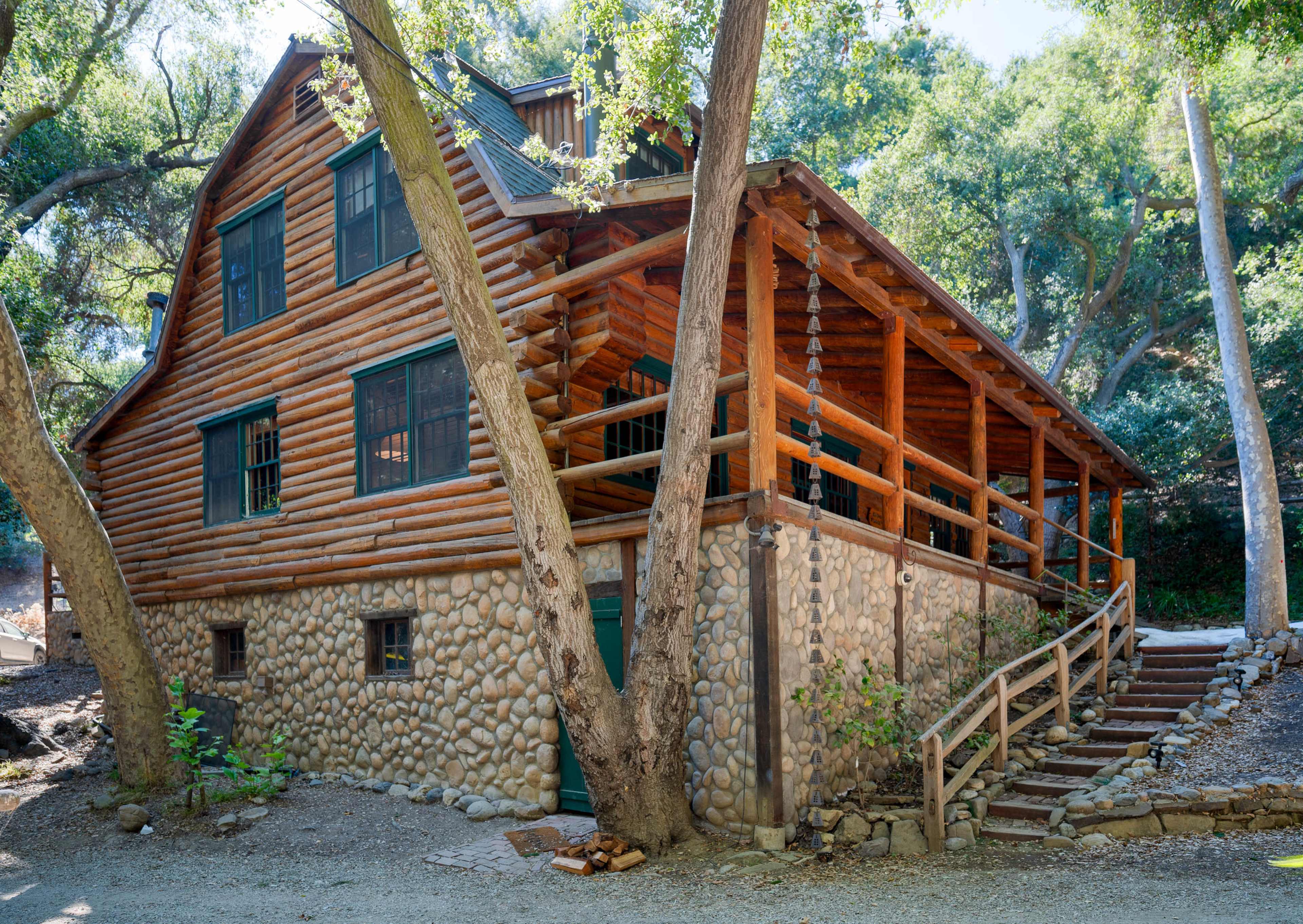 A log cabin with a stone foundation and a wraparound porch is surrounded by trees and situated on a gravel path.