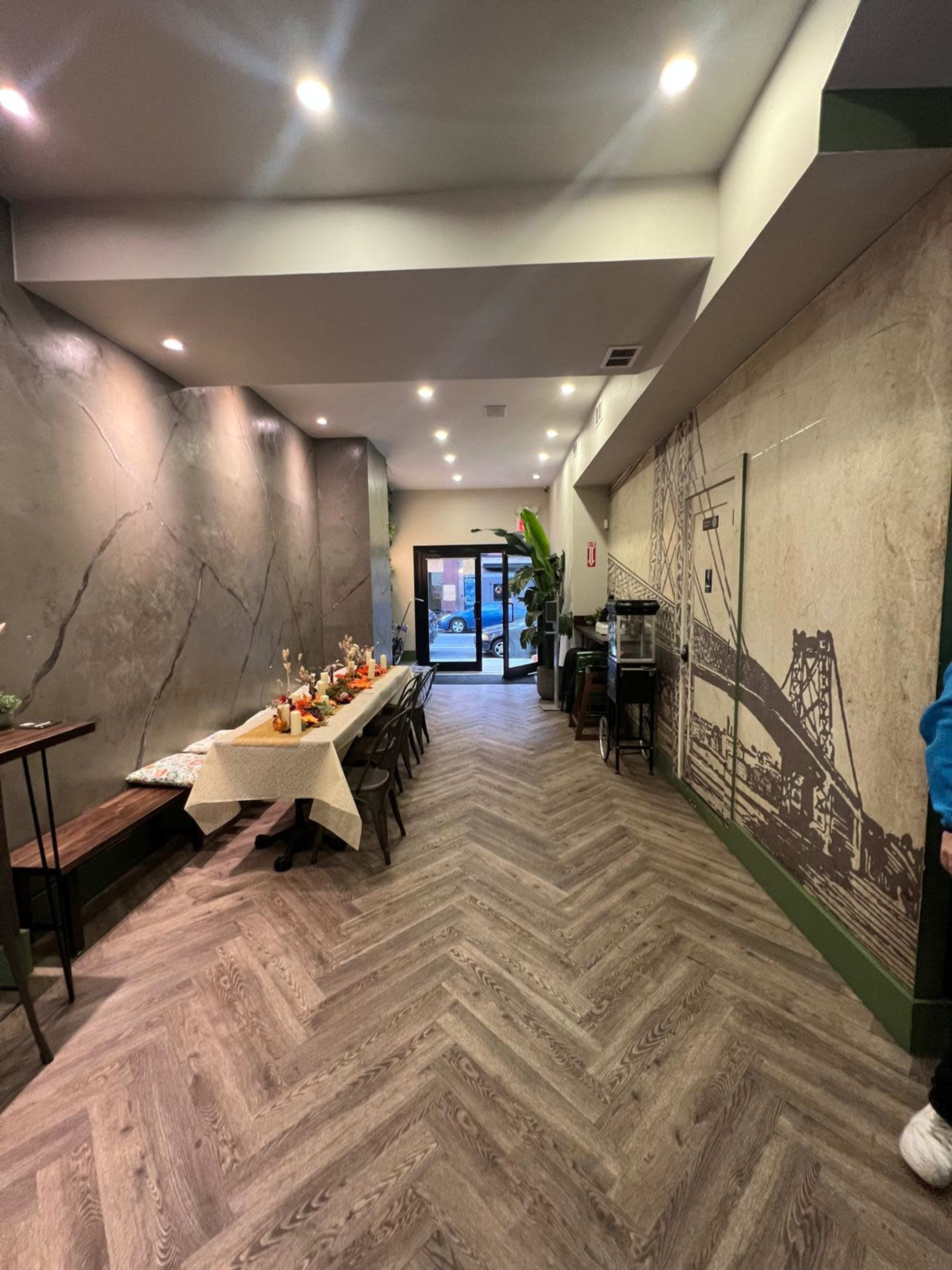 The image shows a spacious hallway in a restaurant, featuring a dining setup with a table decorated for an event, and large windows providing natural light at the entrance.